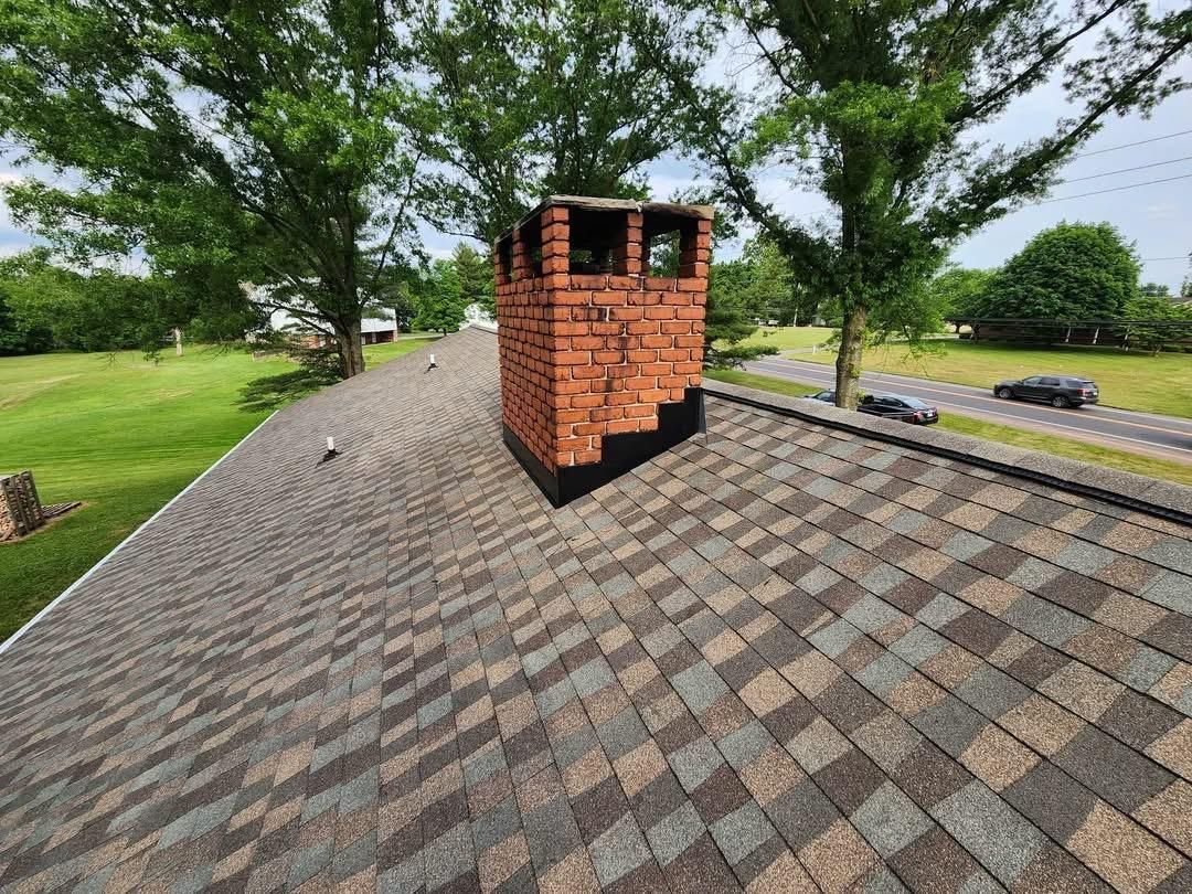 Roof with gray shingles and a brick chimney on a tree-lined roadside.