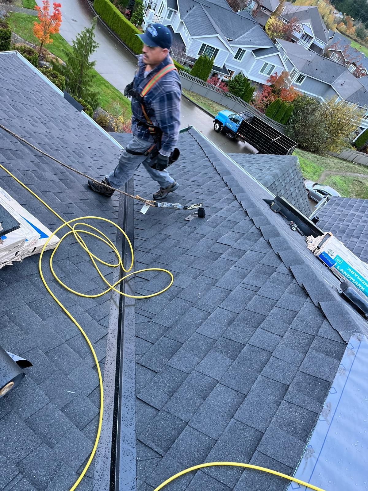 Worker on a steep shingled roof, walking near yellow hoses and tools, with houses in the background.