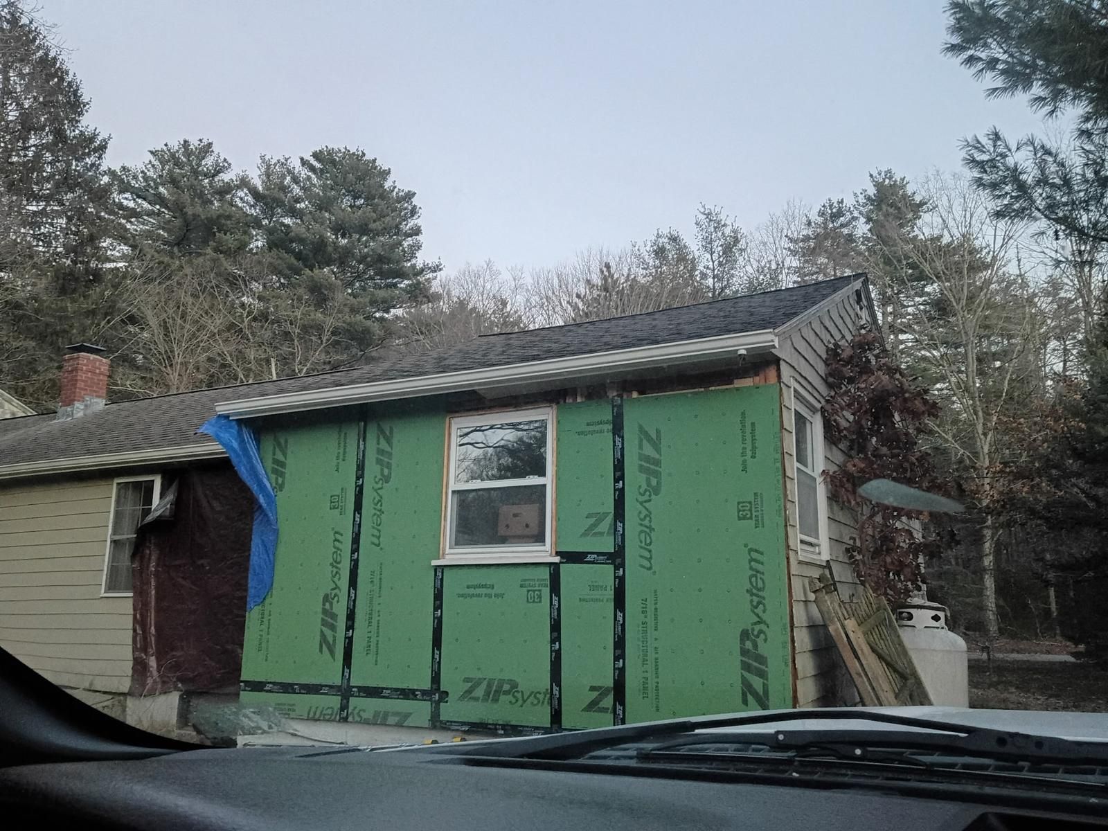 Small house exterior under renovation, with green house wrap and a window, beside trees and a parked vehicle