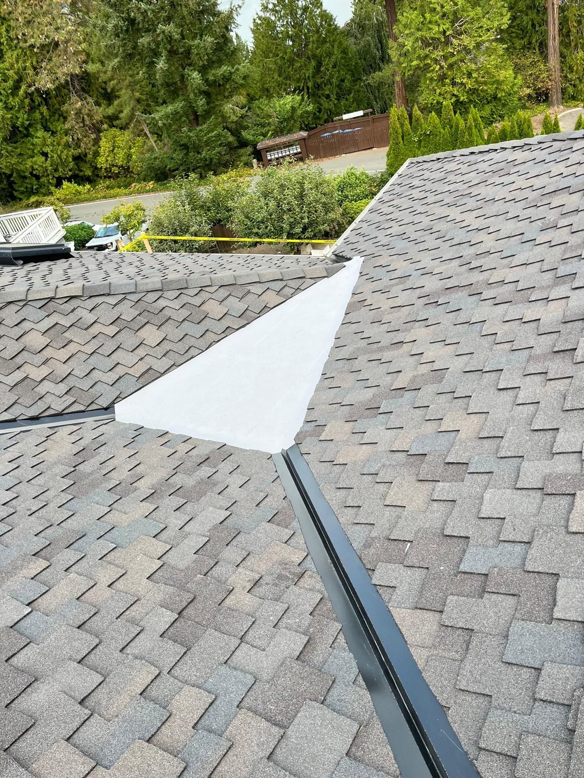 Gray shingle roof with a torn white tarp over a seam, with trees in the background