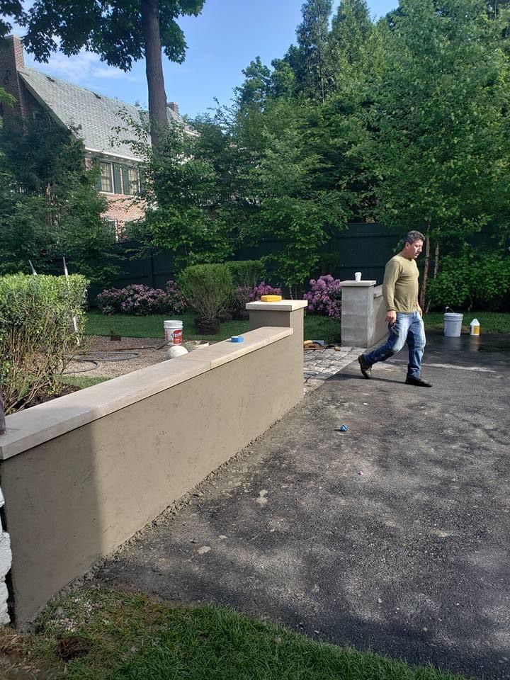 Person walking on a paved garden path beside a beige wall, with trees and flowers in the background.