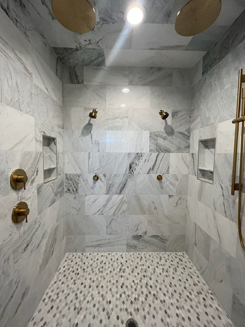 Marble-tiled shower with multiple gold fixtures and pebble-pattern floor in a bright bathroom