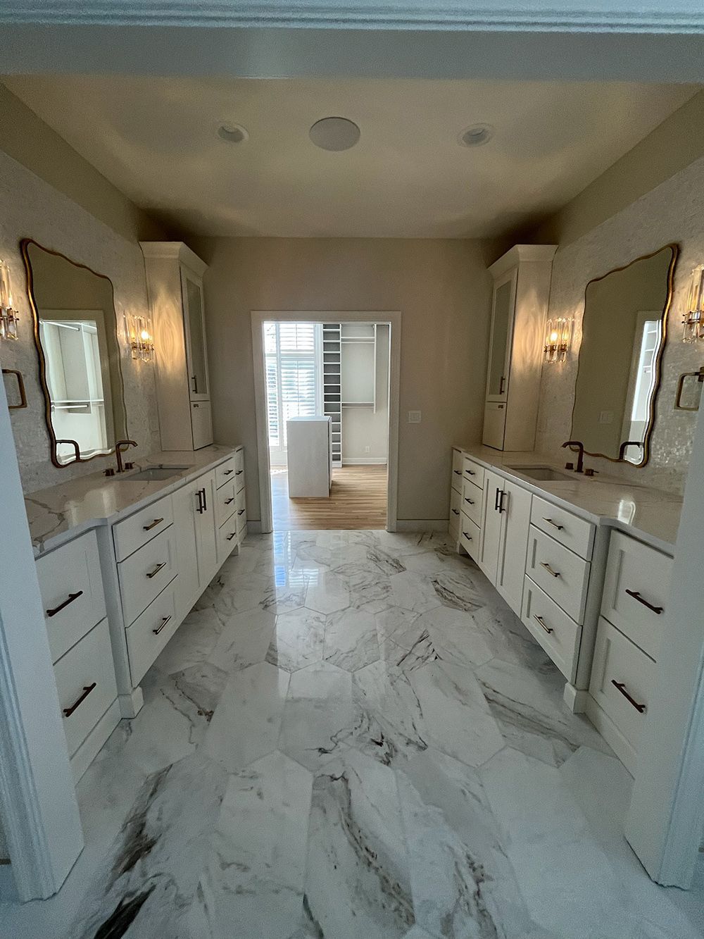 Narrow luxury bathroom with double vanities, white marble flooring, and a doorway to another room.