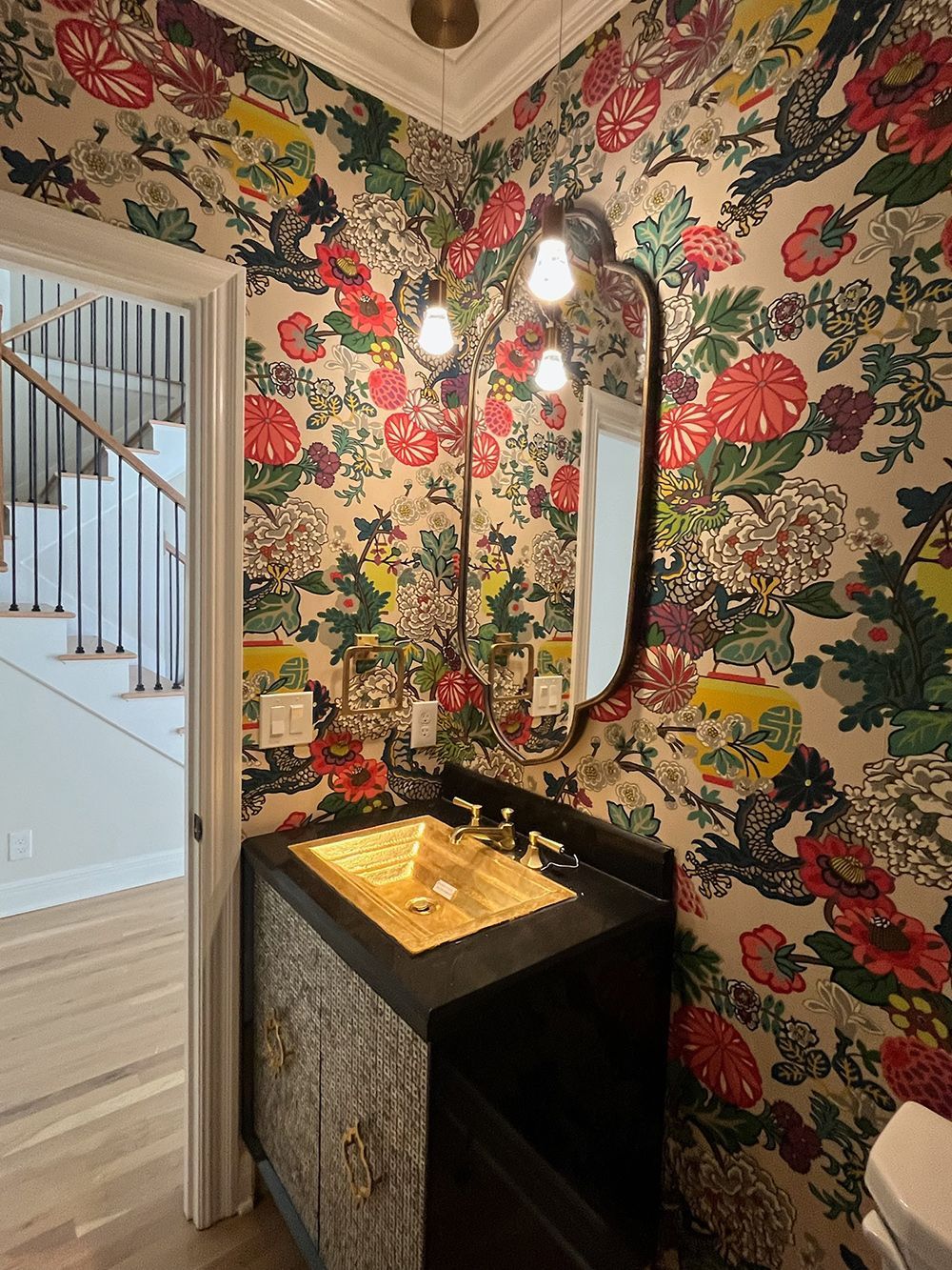 Floral wallpaper bathroom with black vanity, gold sink, and oval mirror by an open doorway