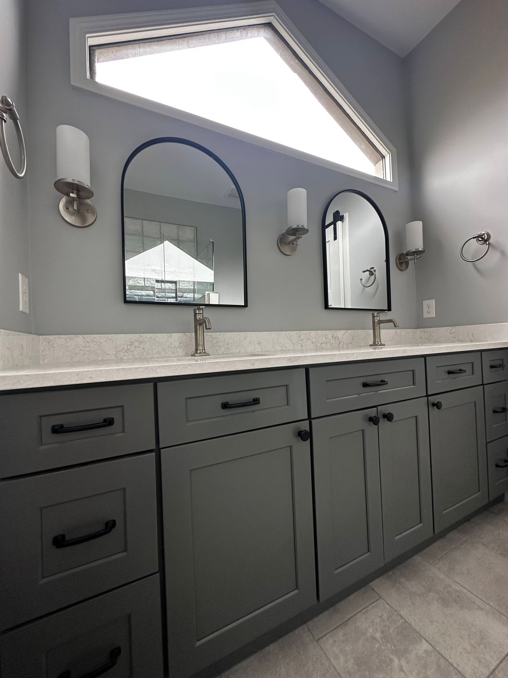 Bathroom with gray cabinets, mirrors, sconces, and a triangular window.