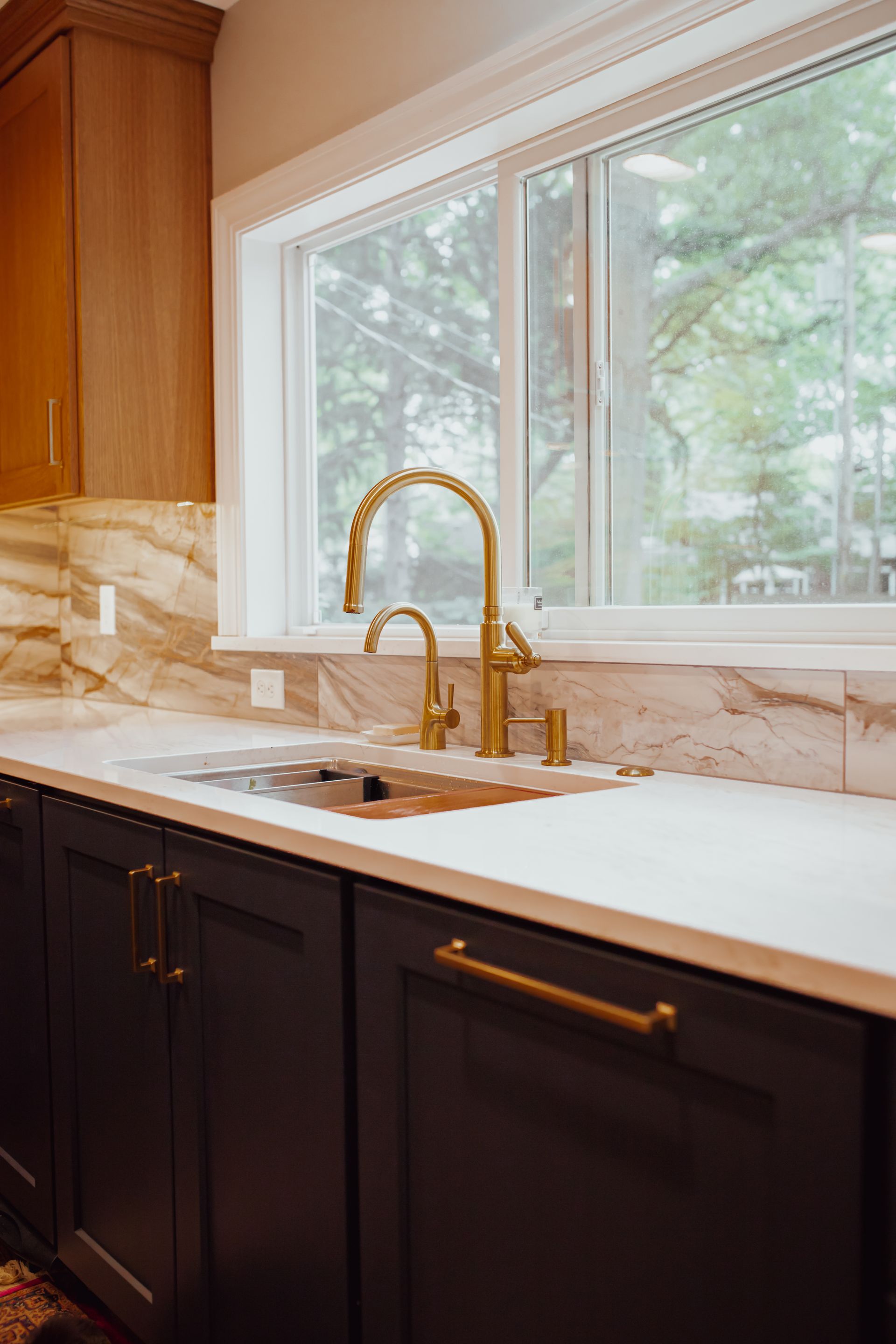 Kitchen with dark cabinets, gold faucet, marble backsplash, and a window.