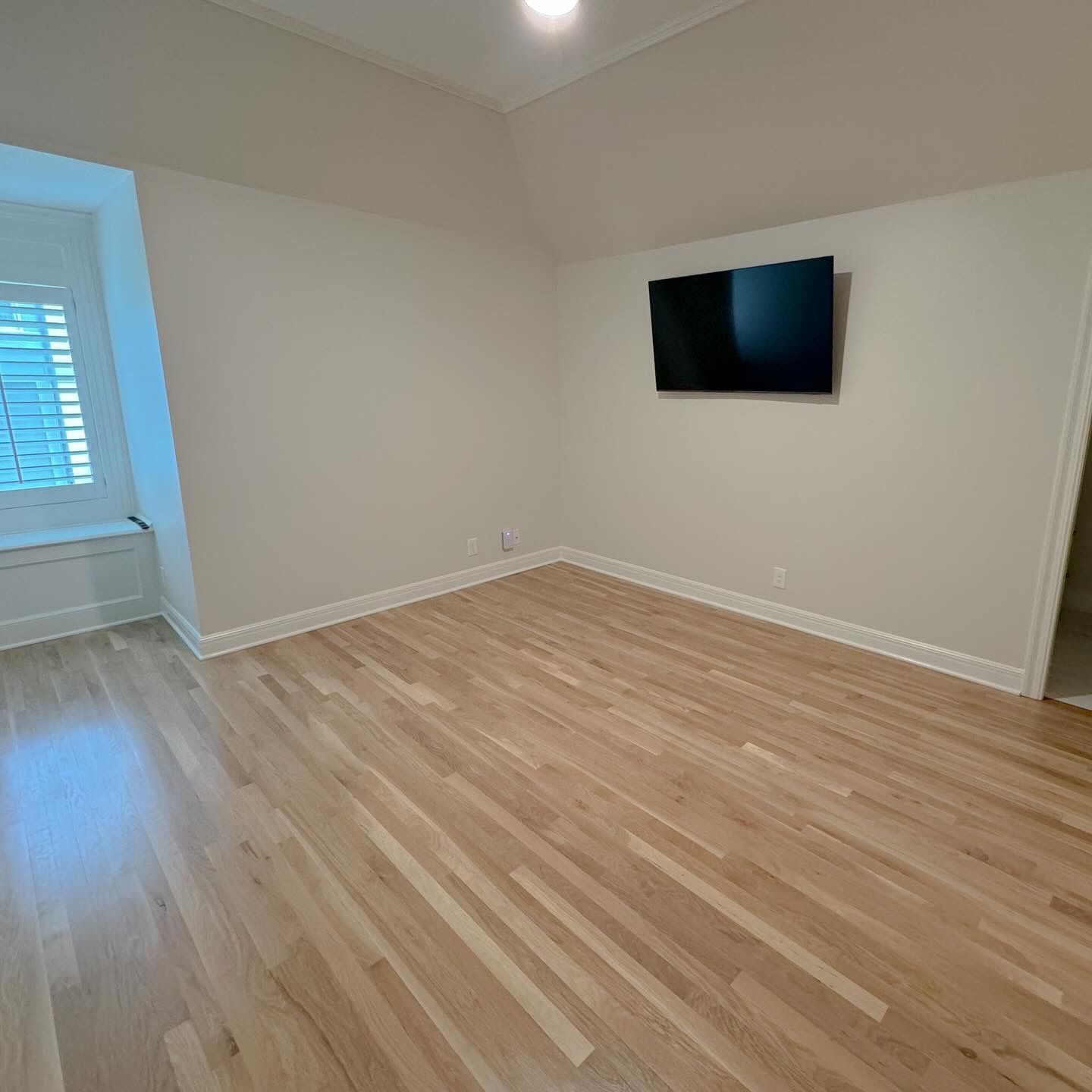 Empty room with hardwood floors, a mounted TV, and a window with shutters