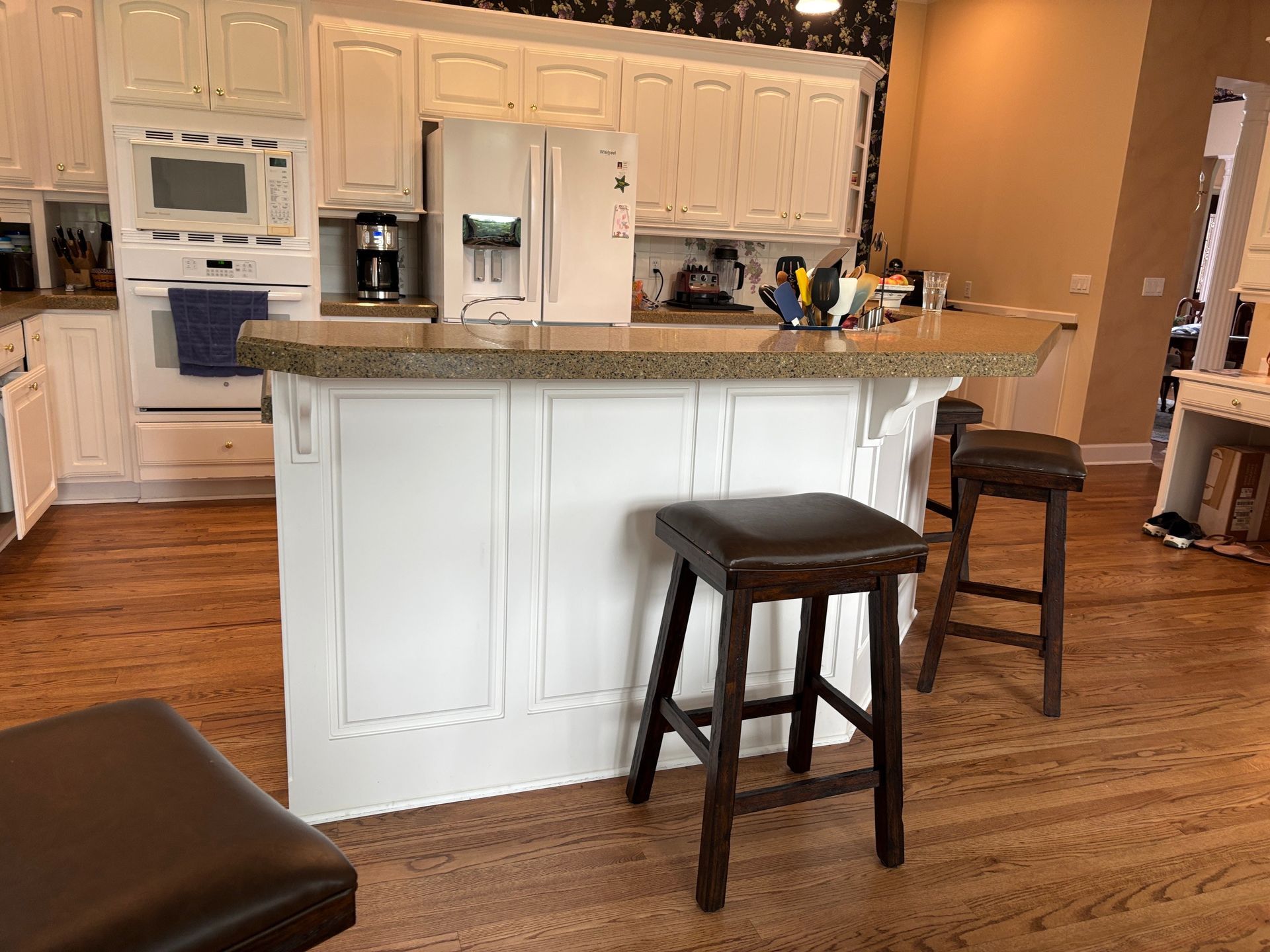 A kitchen features a white island with a speckled countertop and two dark stools on a wood floor, facing white cabinets.