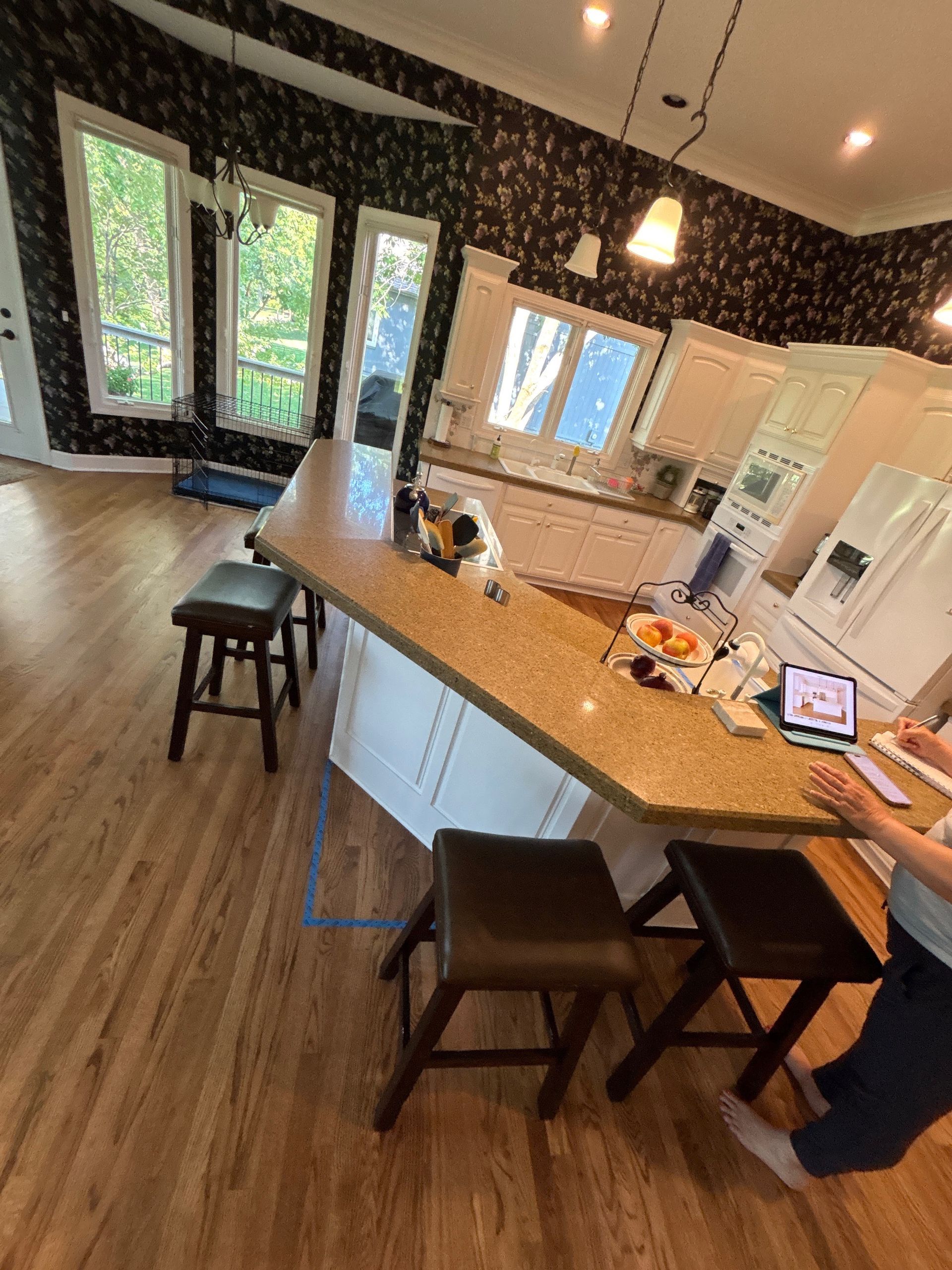 A bright kitchen with white cabinetry, a tan granite countertop peninsula, three bar stools, and floral wallpaper.