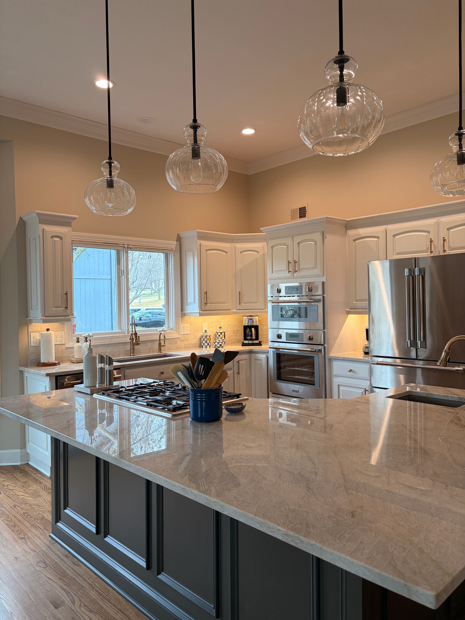 A bright kitchen with a large gray island, granite countertops, white cabinetry, and three hanging glass pendant lights.