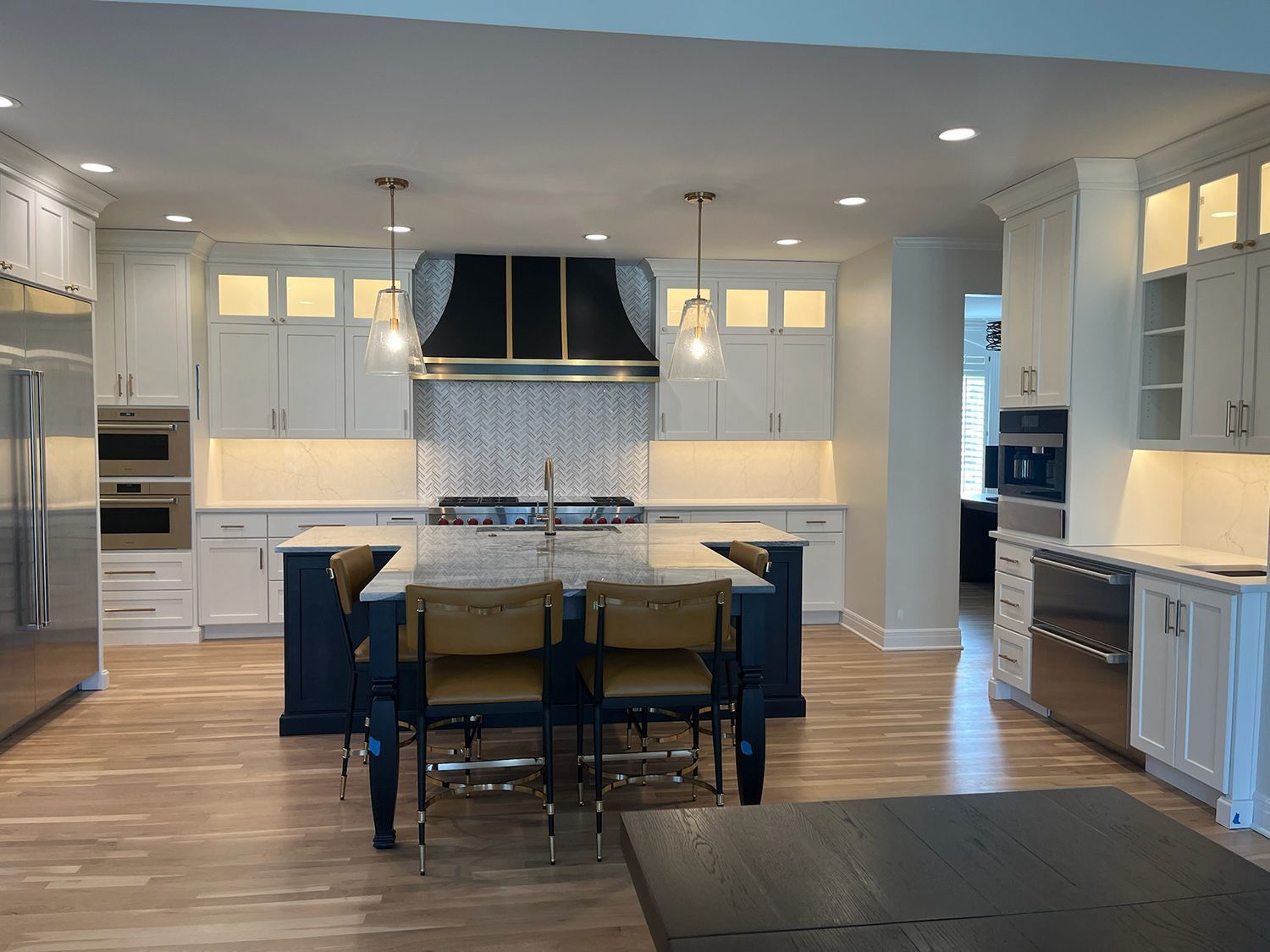 Modern white kitchen with large island, dark chairs, stainless appliances, and wood floors