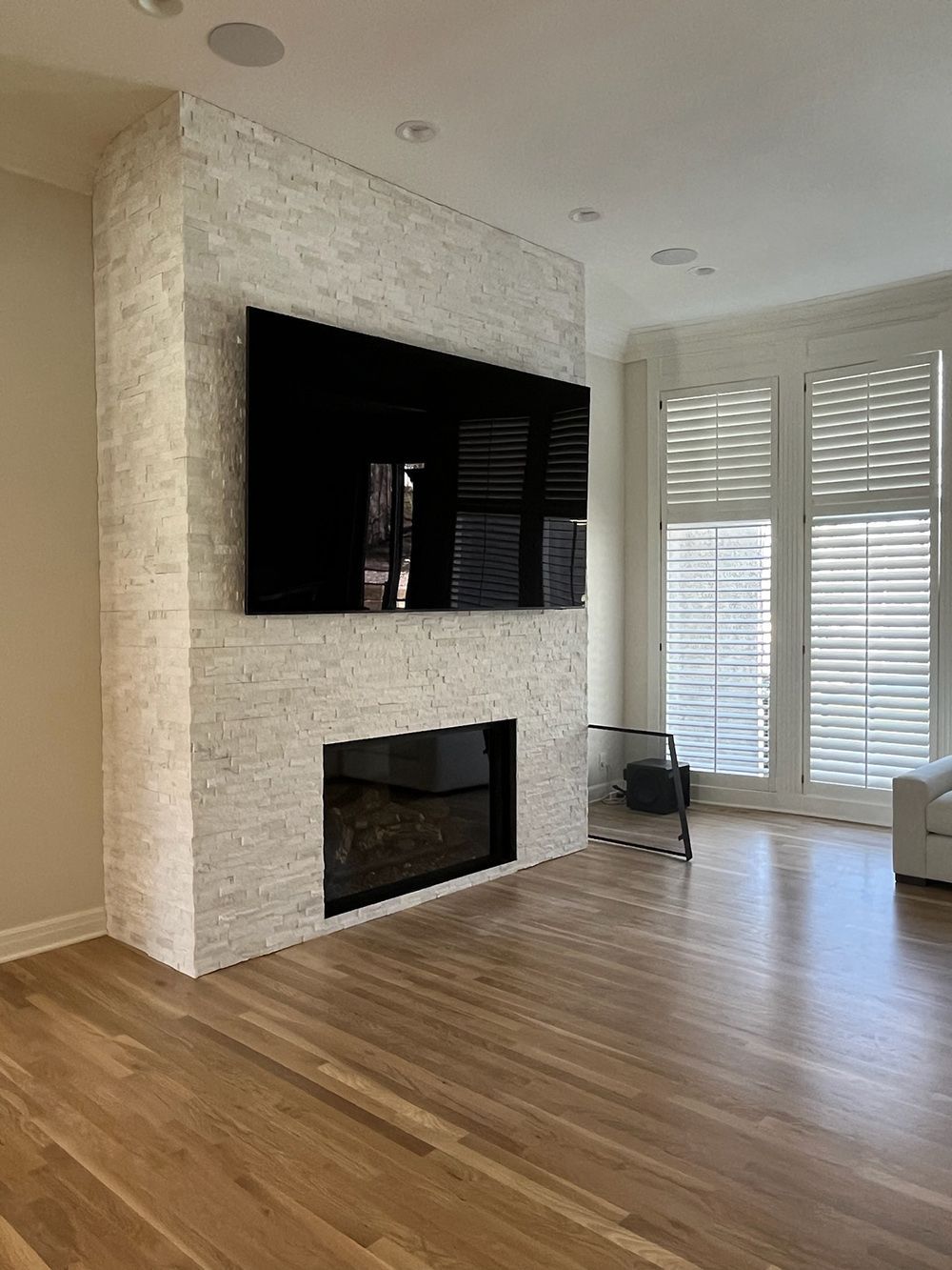 Modern living room with wall-mounted TV, fireplace, wood floor, and large windows with shutters