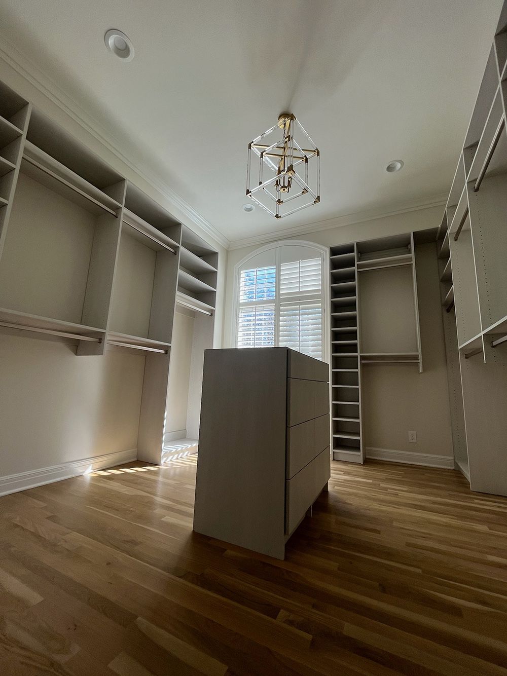 Empty walk-in closet with built-in shelves, hardwood floors, central island, and chandelier under a window.