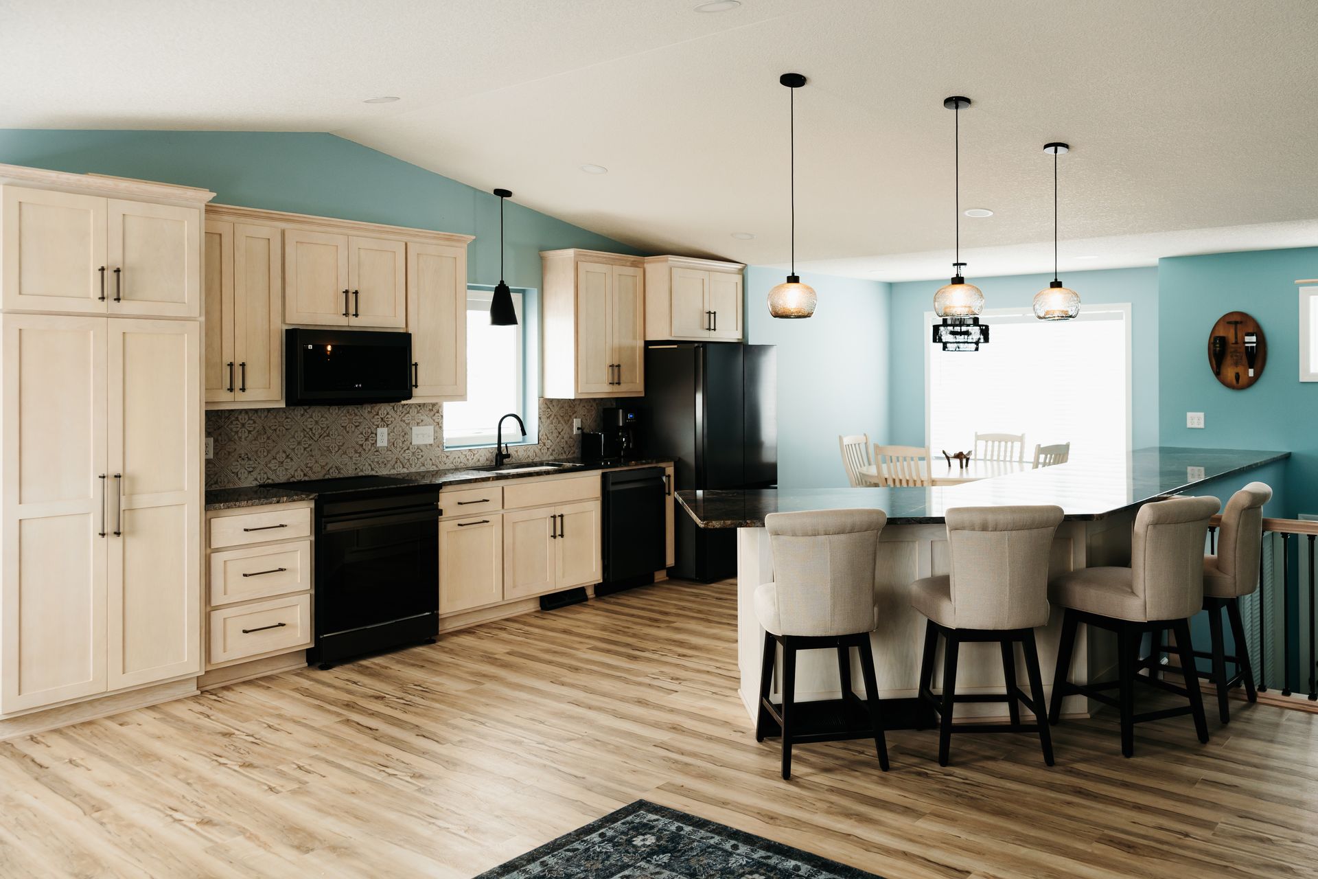 A modern kitchen with light wood floors, cream cabinets, black appliances, and a teal accent wall. A kitchen island has seating.