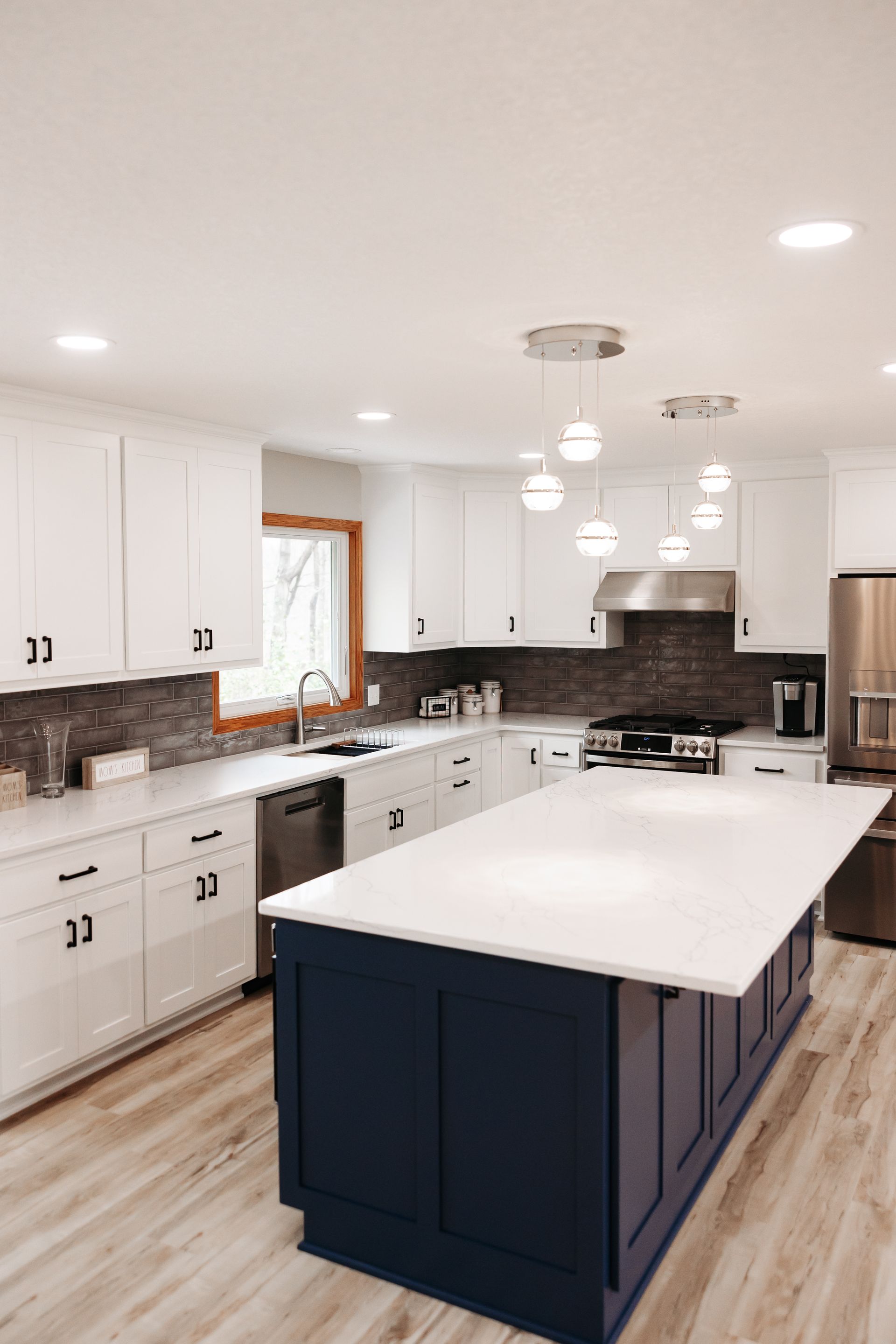 Modern kitchen with white cabinets, navy blue island, and a stainless steel refrigerator. The island has a white countertop with globe pendant lights above it.
