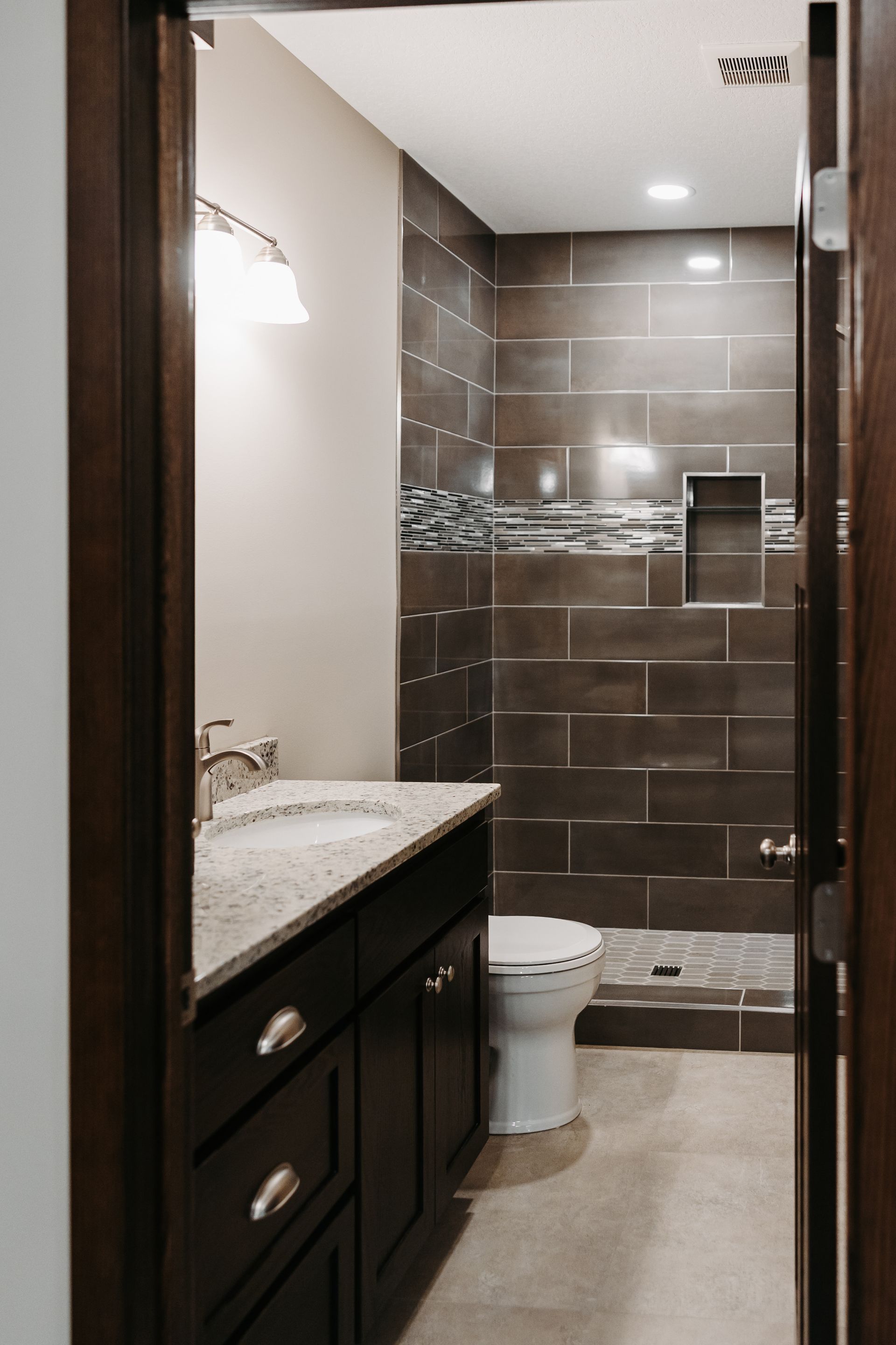 Dark-toned bathroom with dark brown vanity and tile shower. Vanity has a light countertop and sink, with a toilet beside the shower.