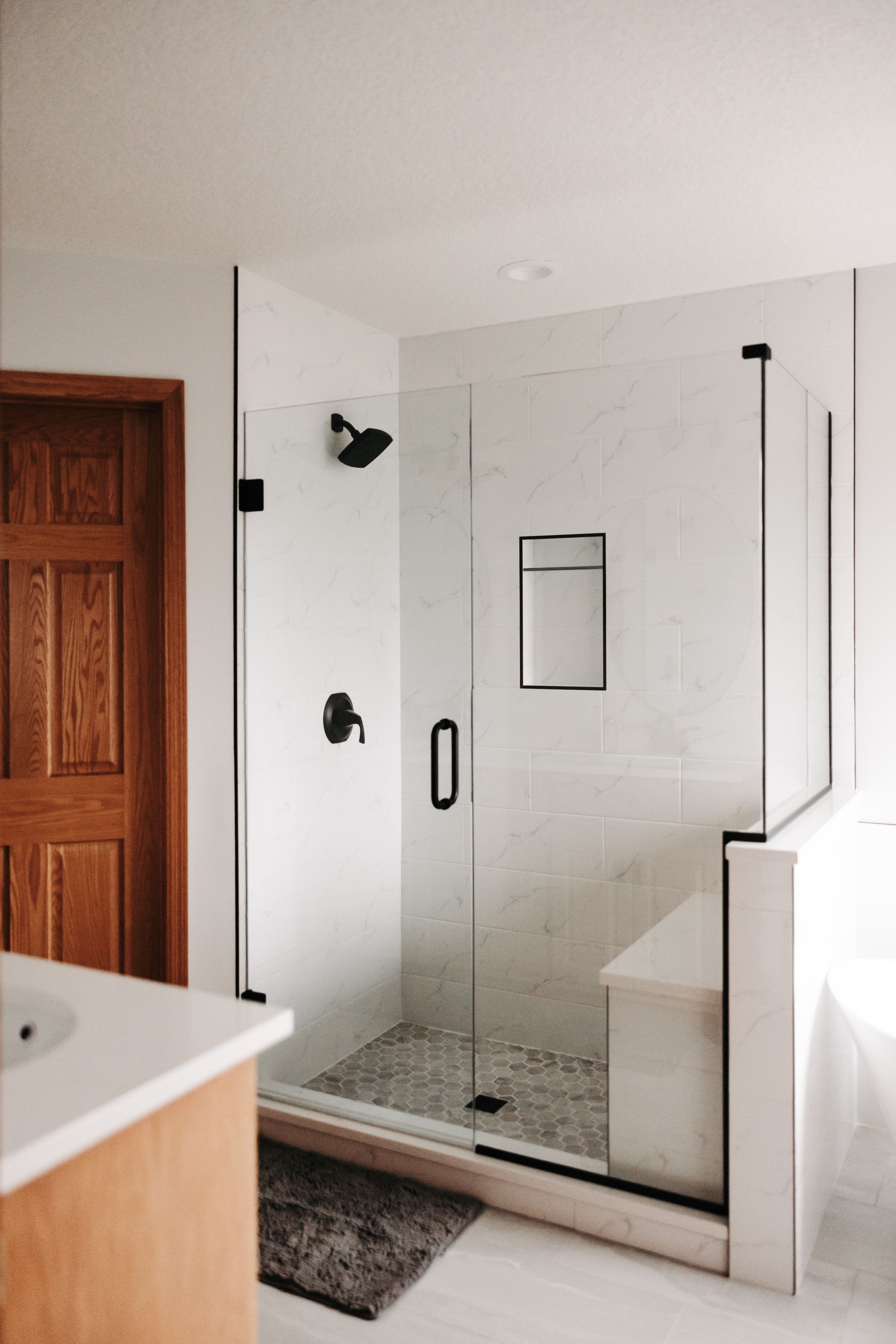 Modern bathroom with glass shower and dark fixtures. White walls, stone floor in shower, wood door.