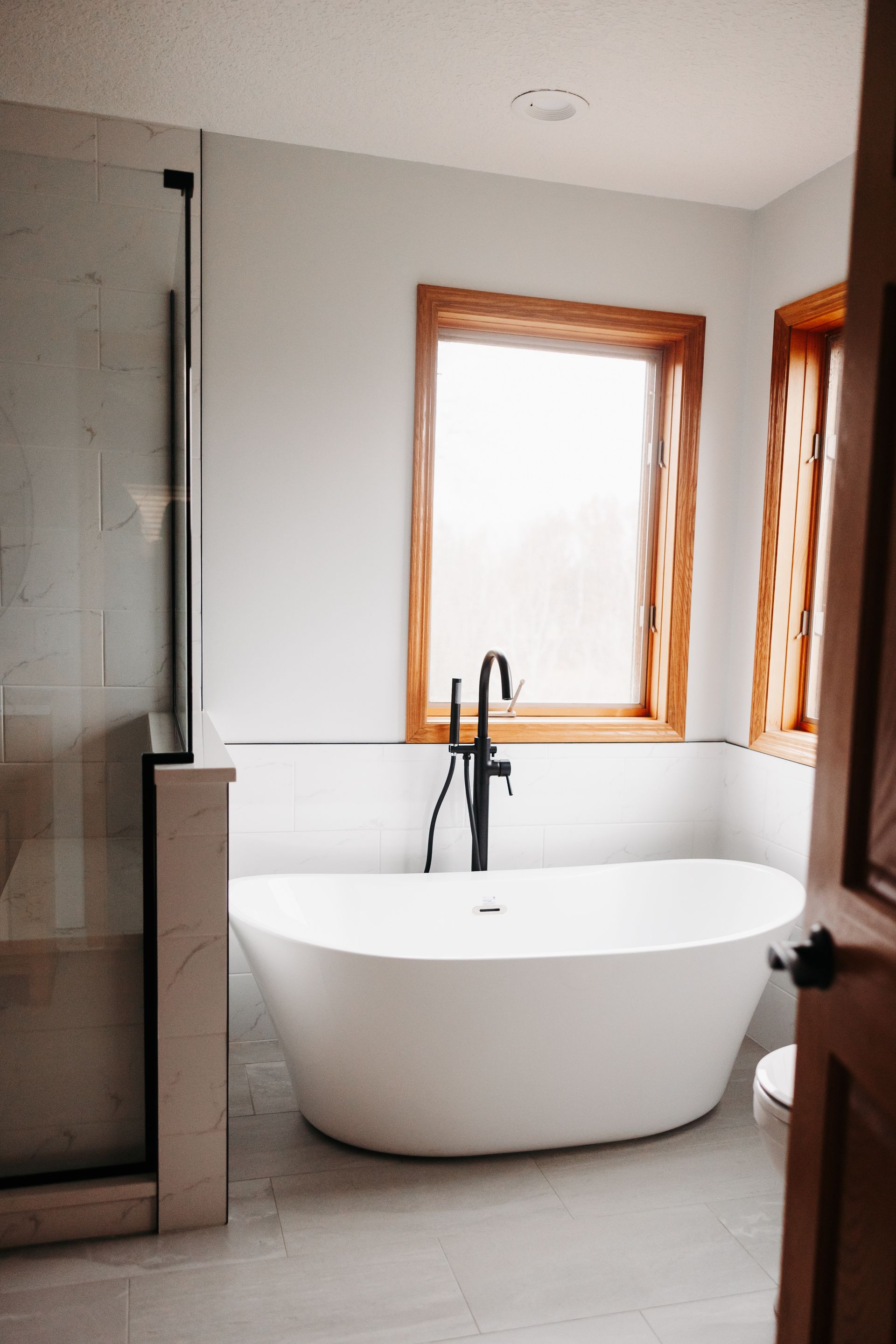 Modern bathroom with a white freestanding tub, black faucet, and large window with wood trim. A glass shower enclosure is visible on the left.