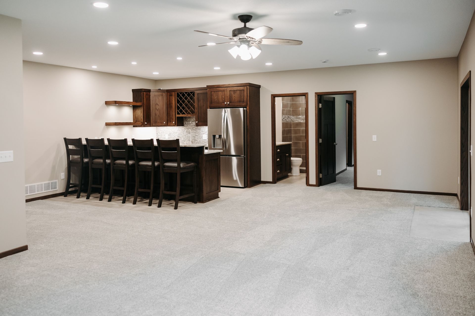 Basement with a bar, dark wood cabinets, stainless steel refrigerator, and a row of bar stools. There are two open doors, and the floor is covered in carpet.