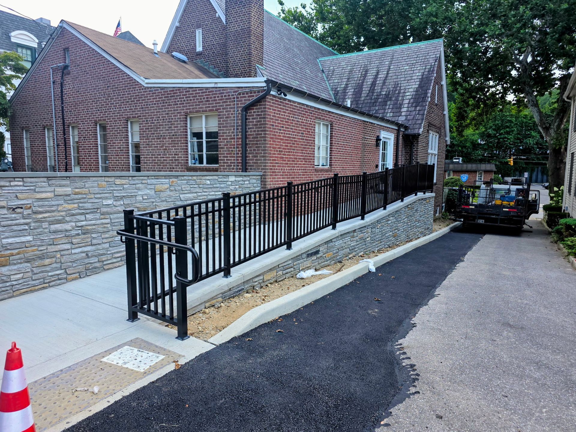 Brick building beside a ramped sidewalk with black railings and a parked car on a narrow street.