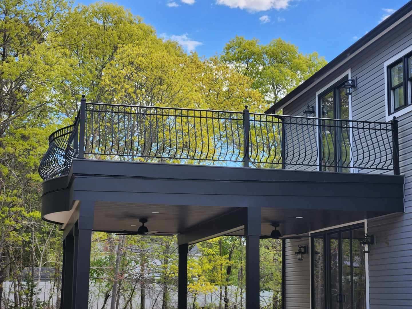 Raised black deck with wrought-iron railings beside a gray house, overlooking trees in spring.