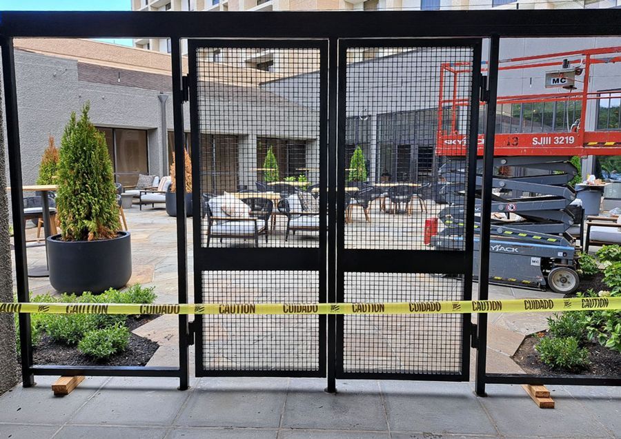 Closed black metal gate blocking a courtyard with potted plants, caution tape, and parked equipment beyond