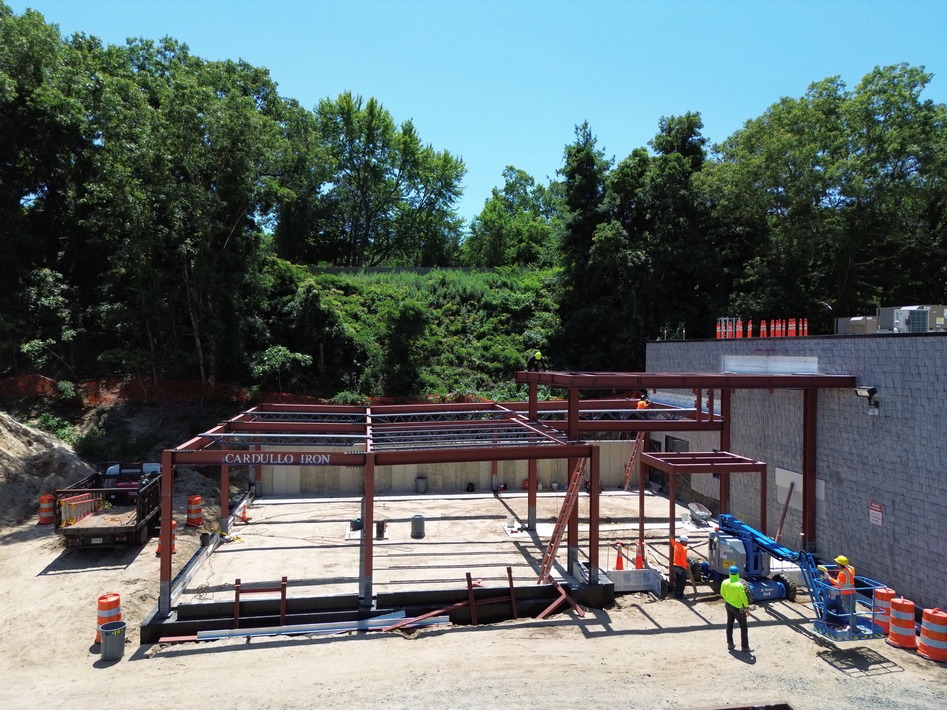 Steel frame building under construction with workers, concrete slab, and trees in the background