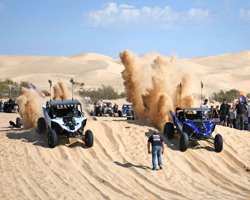 Two off-road buggies race through sand dunes, kicking up large dust clouds, with spectators nearby.