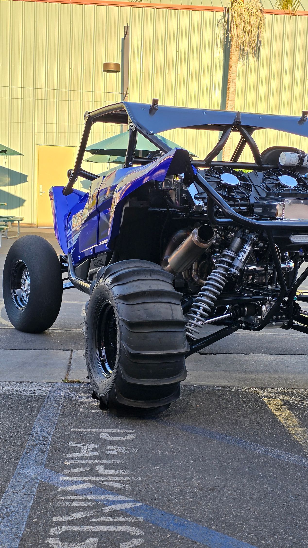 Blue off-road dune buggy with oversized tires parked on pavement beside a yellow wall.