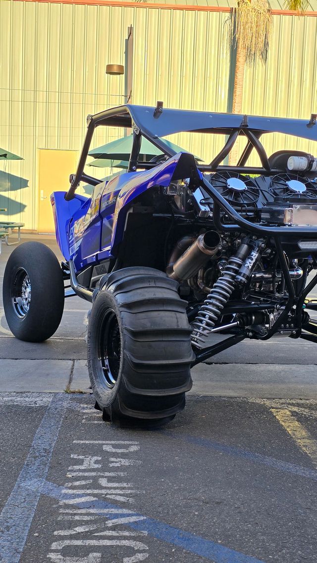 Blue off-road dune buggy with oversized tires parked on pavement beside a yellow wall.