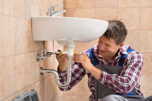 Plumber repairing sink pipes under a bathroom sink, using tools beside a tiled wall.