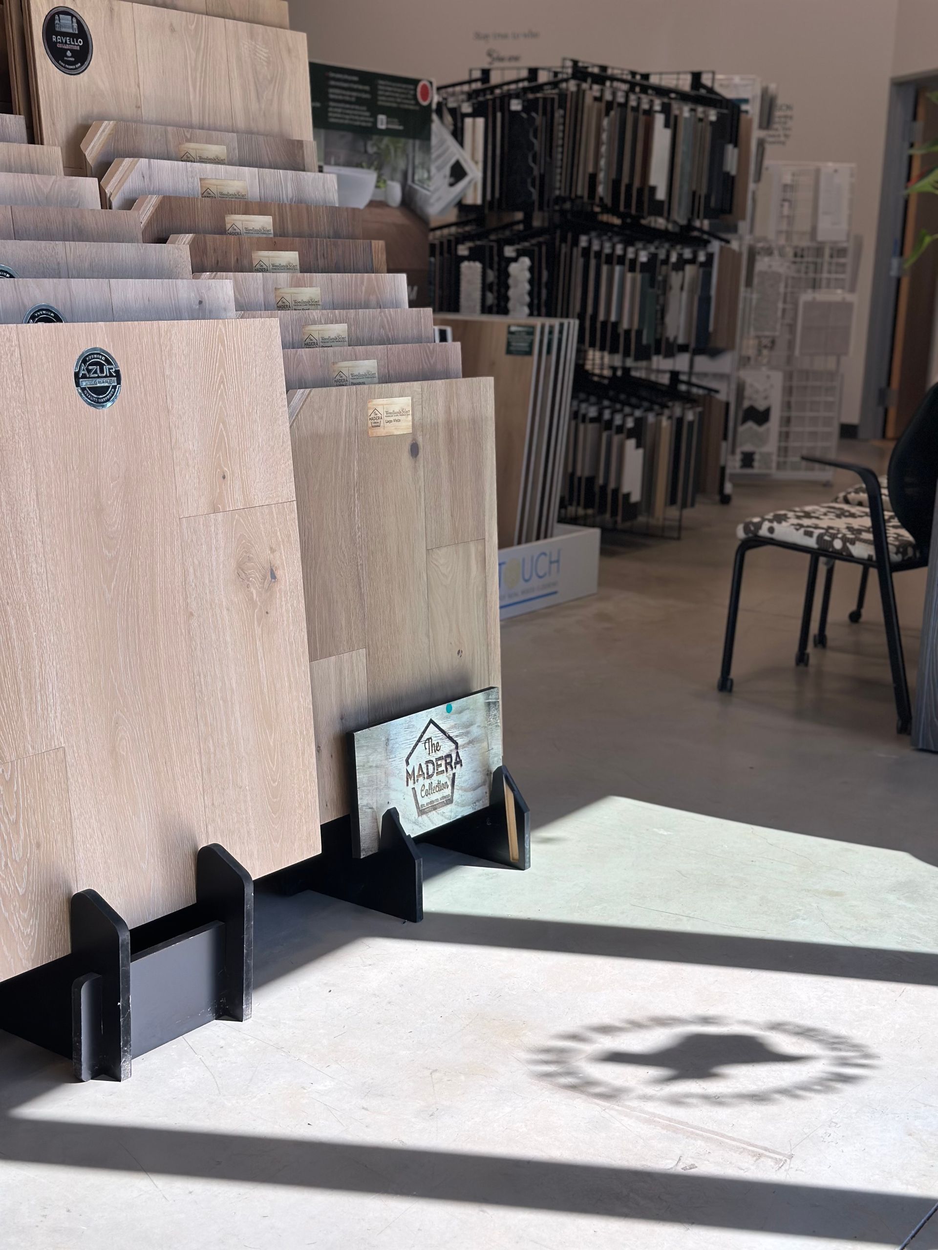 Flooring samples displayed in a showroom. Light wood tones dominate. Shelves with fabric swatches and a chair are visible.