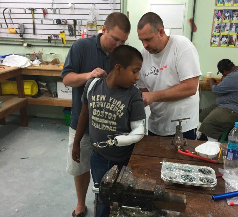 Two men fitting a prosthetic arm on a young boy in a workshop. The boy wears a gray shirt.