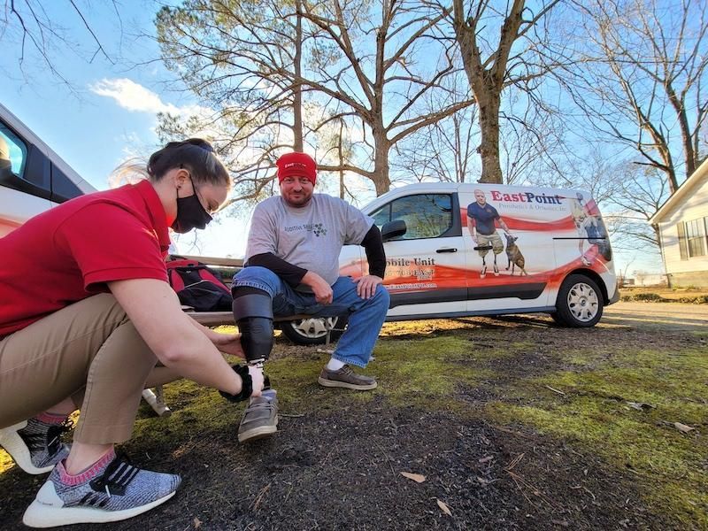 Woman adjusting prosthetic leg on man sitting next to van with a service dog graphic. Outdoor setting.