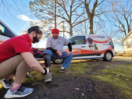 A woman adjusts a prosthetic leg for a man outside a van. The man wears a red hat.