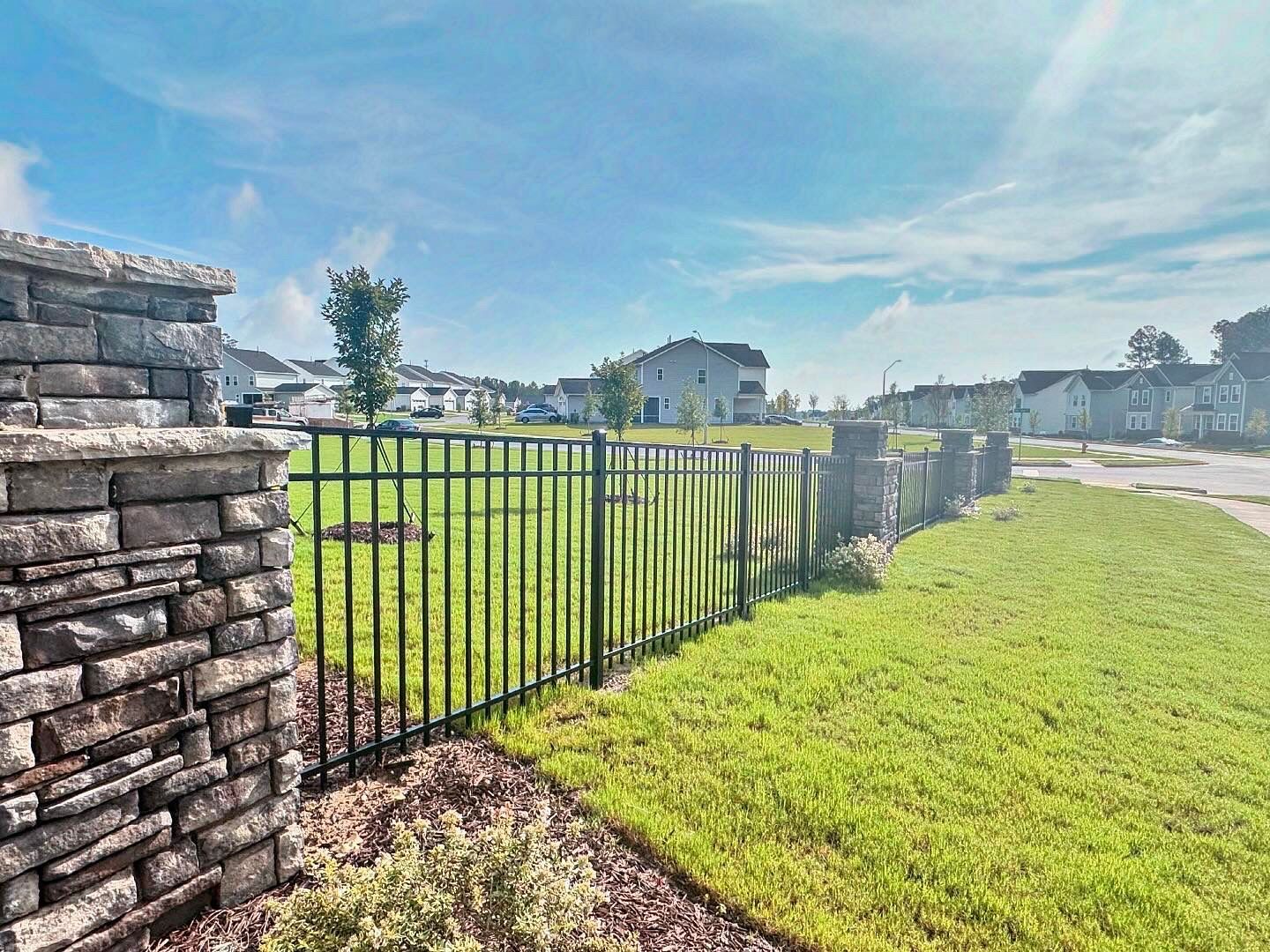 Stone pillars with black metal fence on a grassy lawn; houses and blue sky in the background.