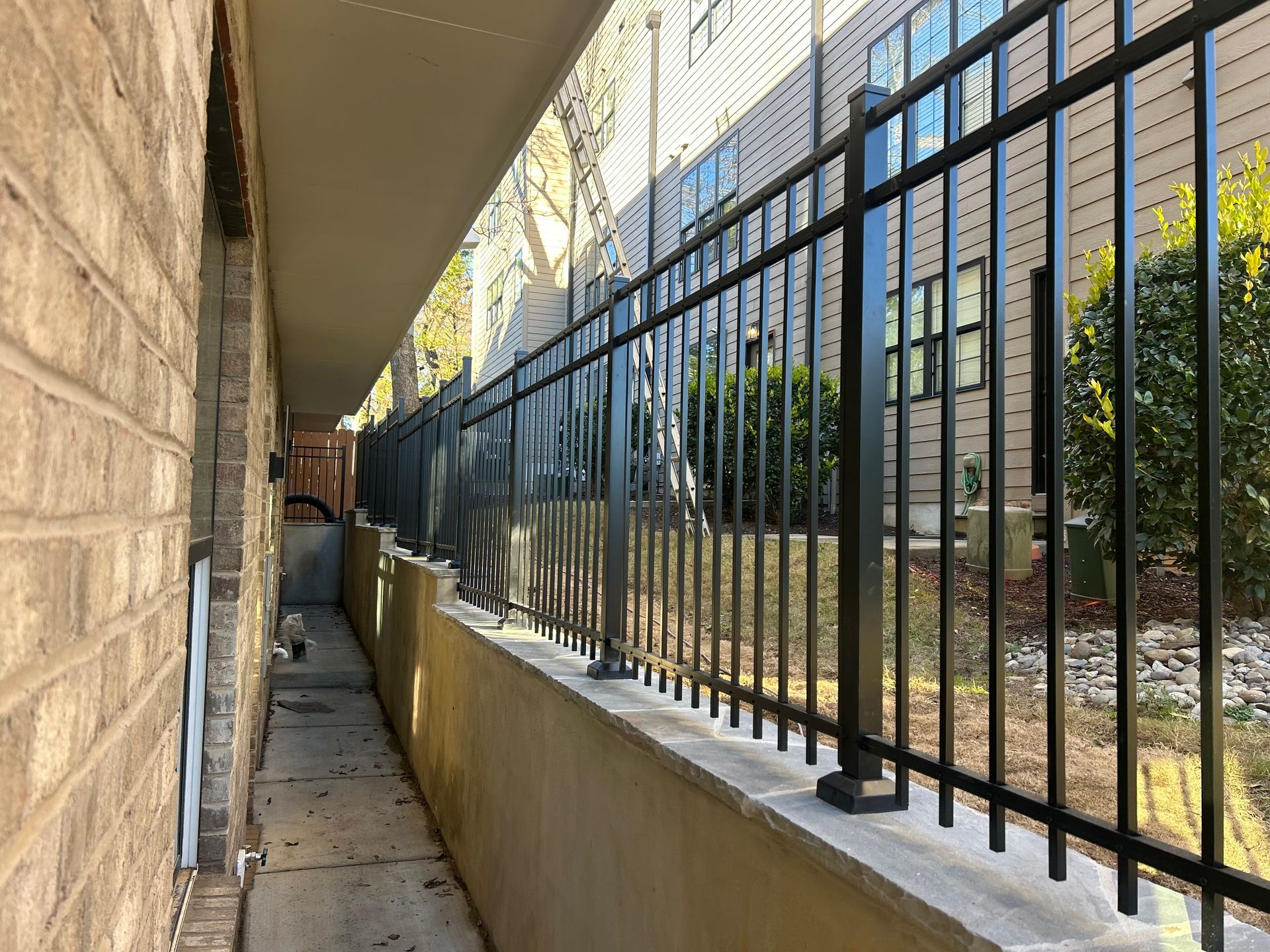 Black metal fence atop a concrete wall along a narrow walkway beside a brick building.