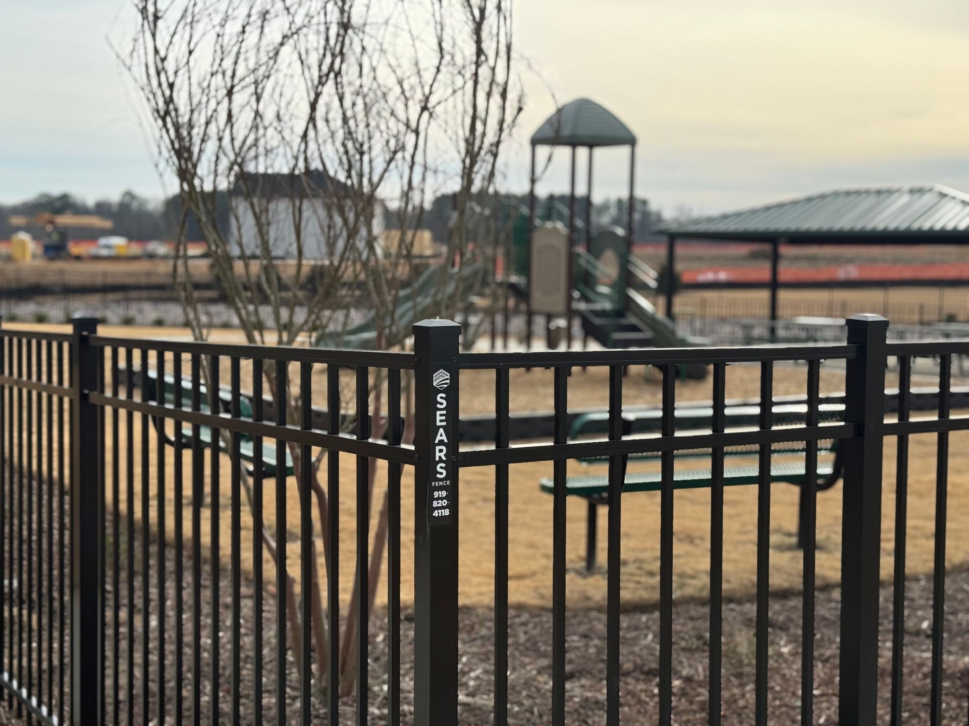 Black fence in front of playground with benches and shelter. Blurred background includes trees and buildings.