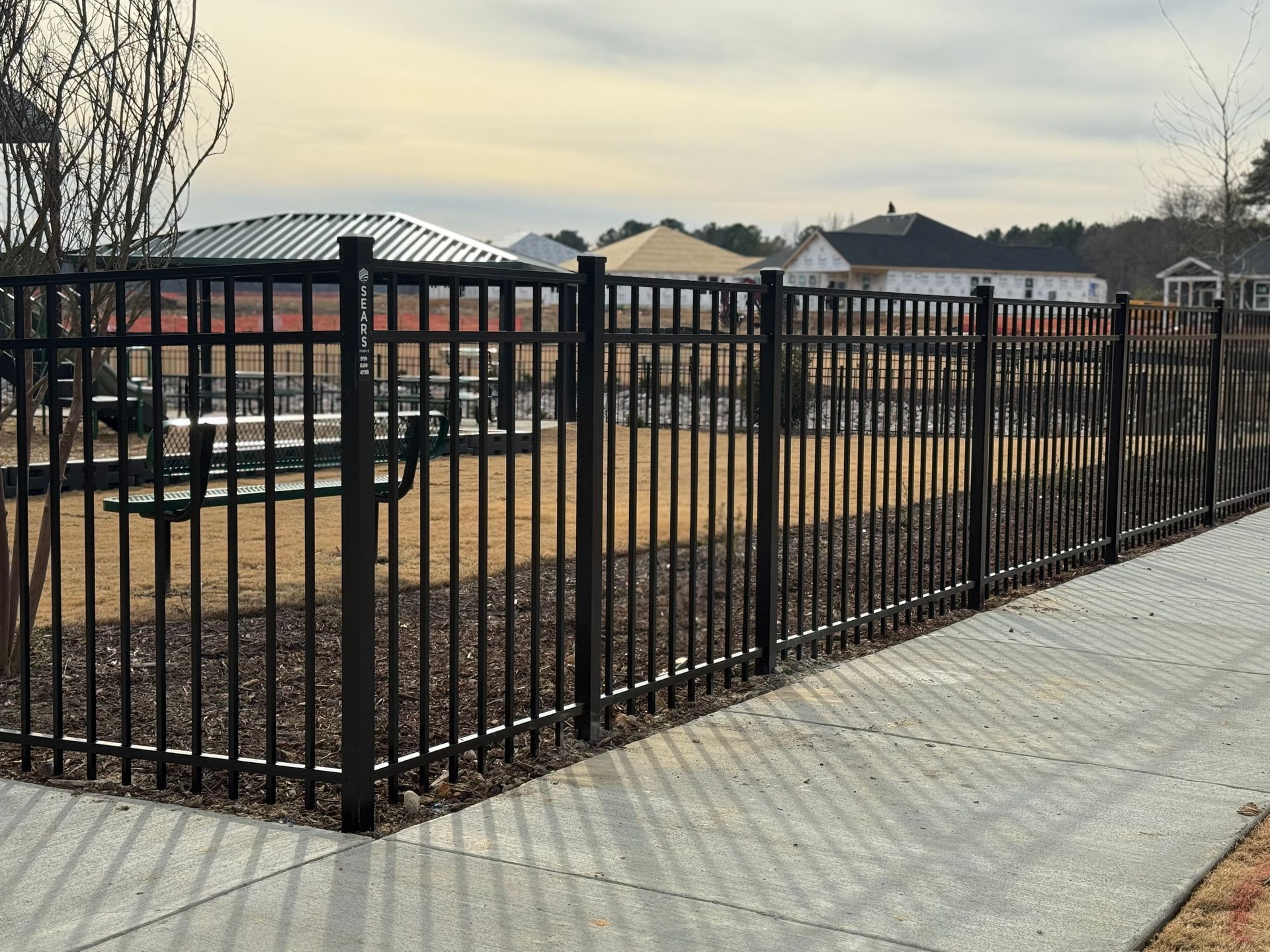 Black metal fence enclosing a park with tables, a pavilion, and houses in the background. Paved path in foreground.