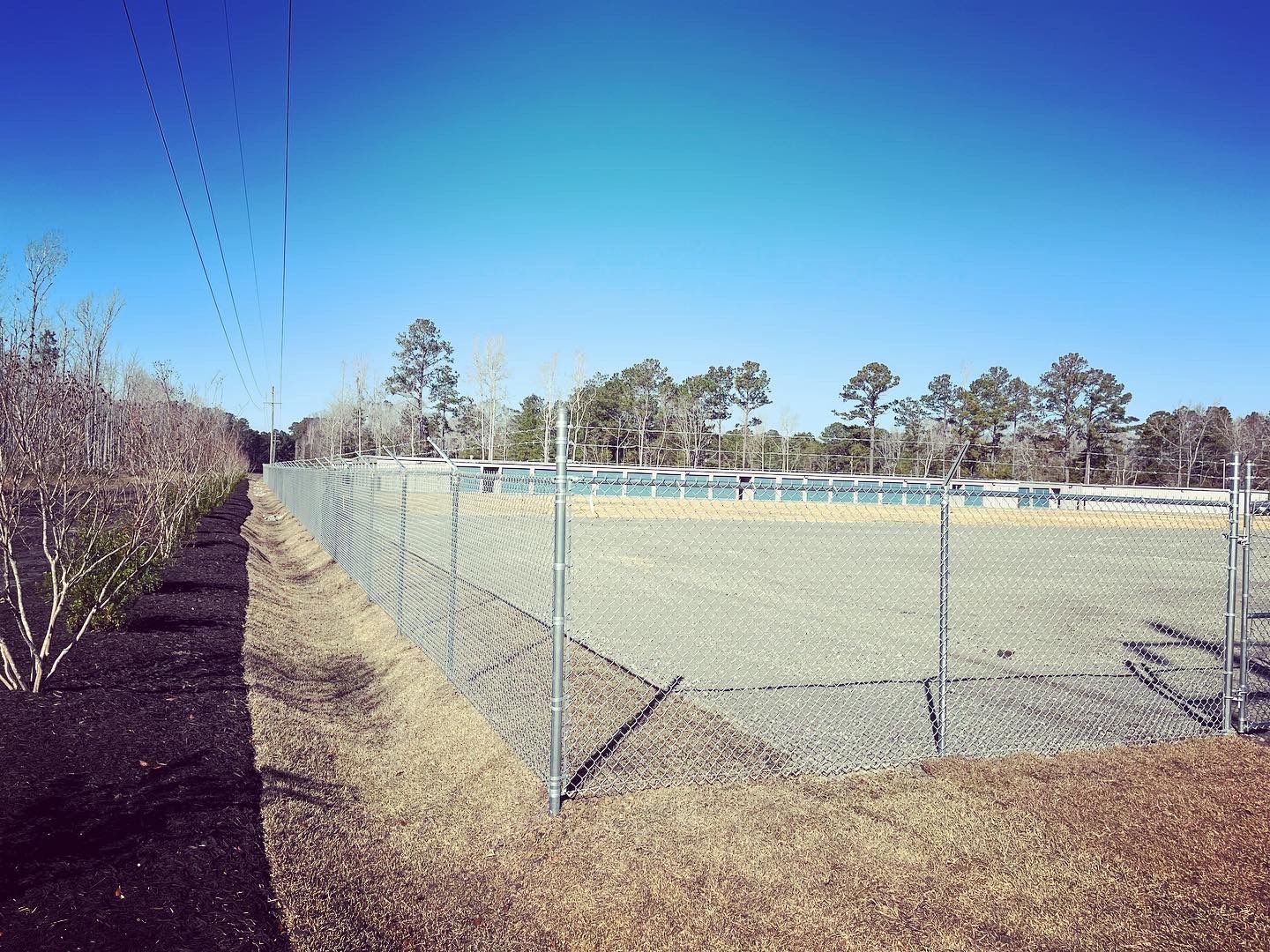 Chain-link fence surrounding an empty field under a clear blue sky, with trees in the background.
