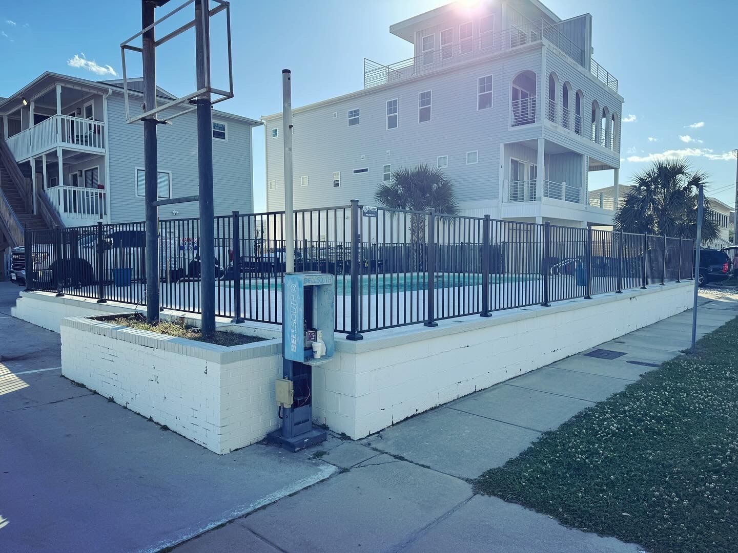 Pool enclosed by black fence, white buildings in background. Bright sunlight.