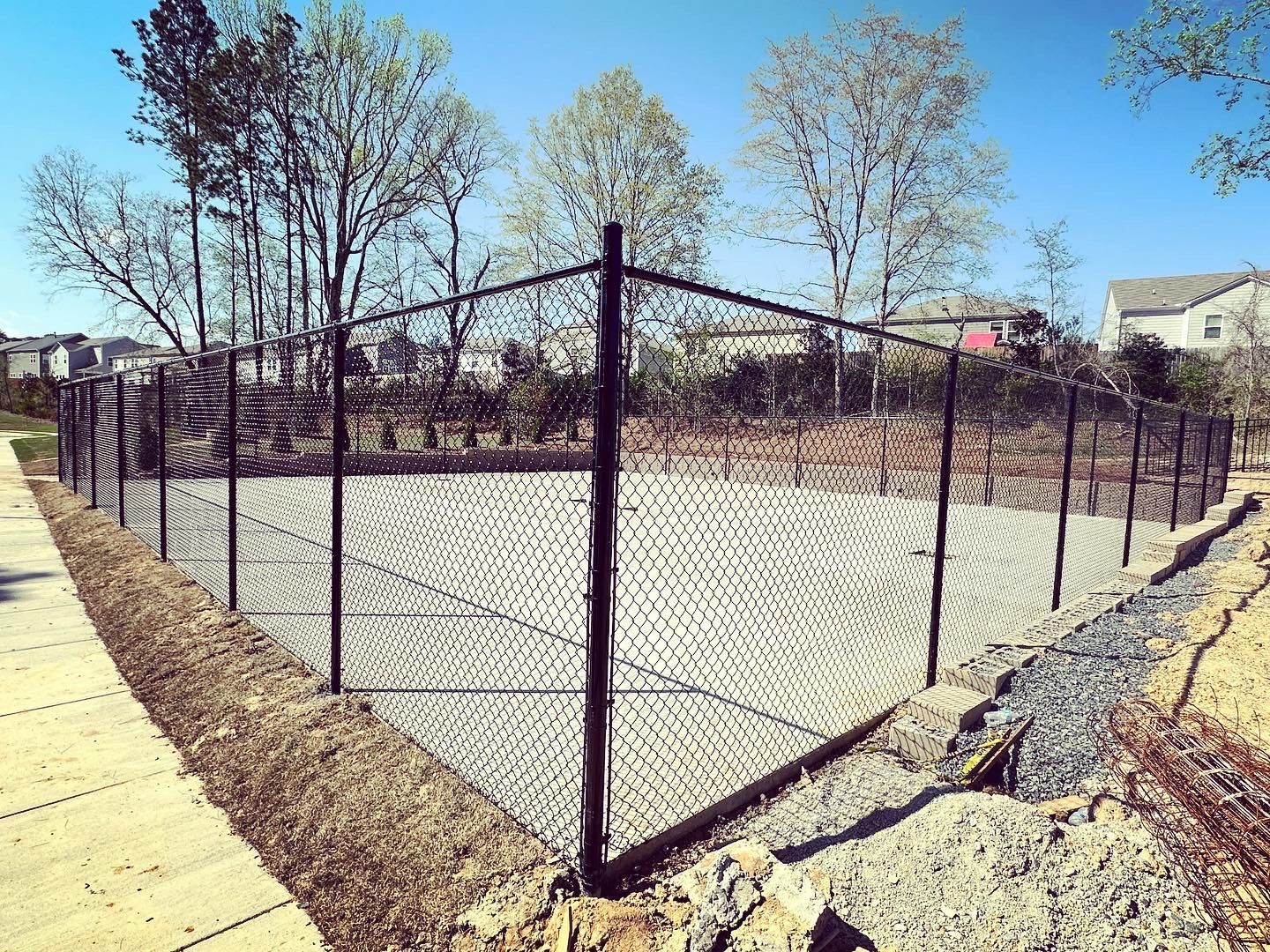 Black chain-link fence surrounds a gravel area; bordering a concrete sidewalk and earth bank, with trees and houses in the background.