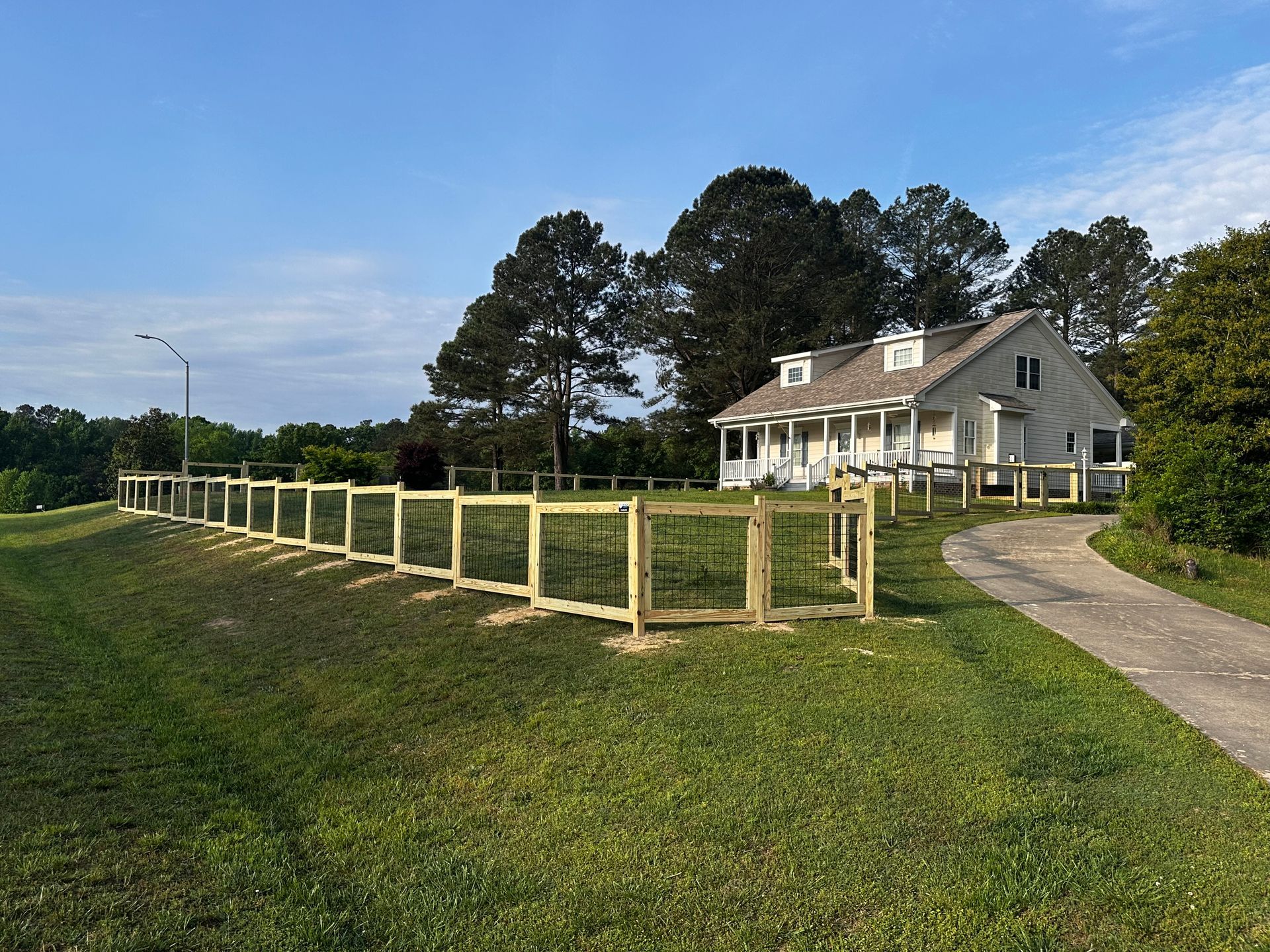 Wooden fence surrounds a white house with a driveway on a grassy hill, under a blue sky.