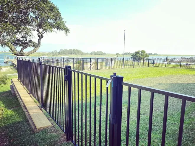 Black fence borders green lawn by calm lake and trees under a blue sky.