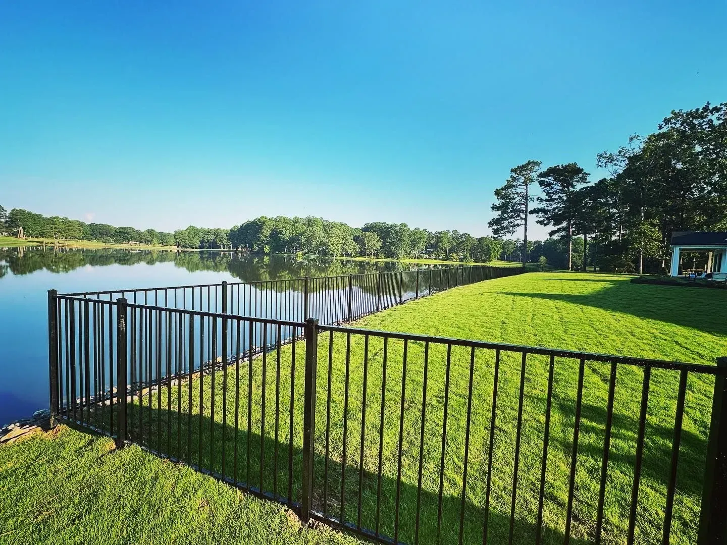Black fence bordering a green lawn next to a lake under a clear blue sky.