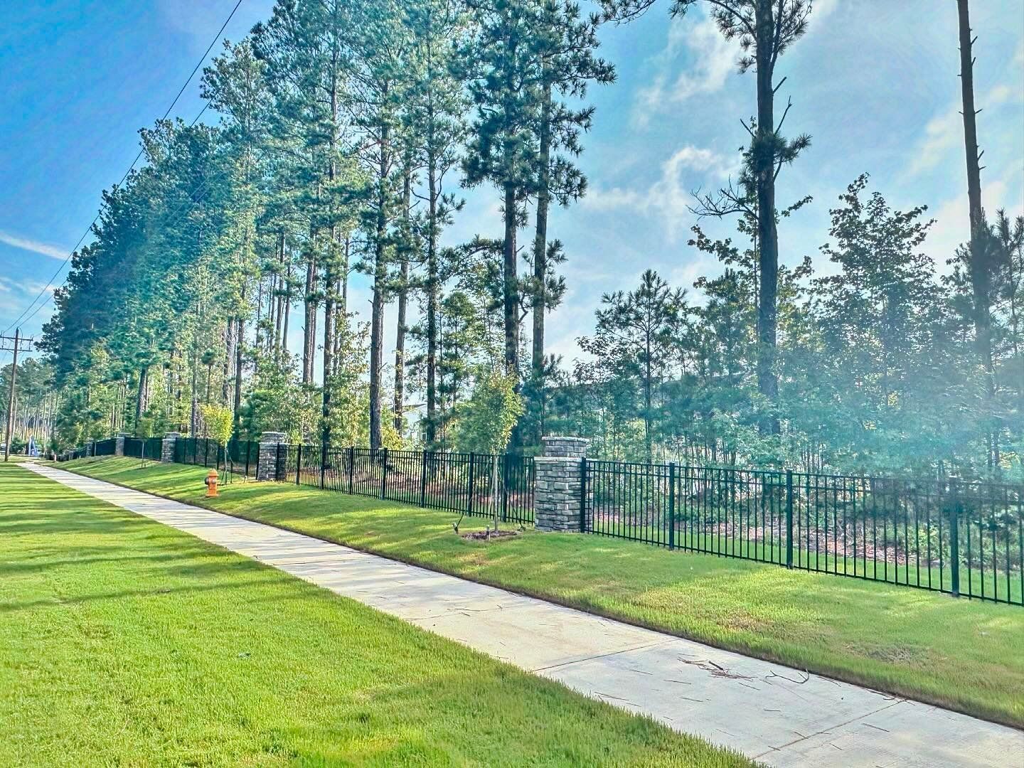 Sidewalk next to a grassy lawn and fence with tall trees under a partly cloudy sky.