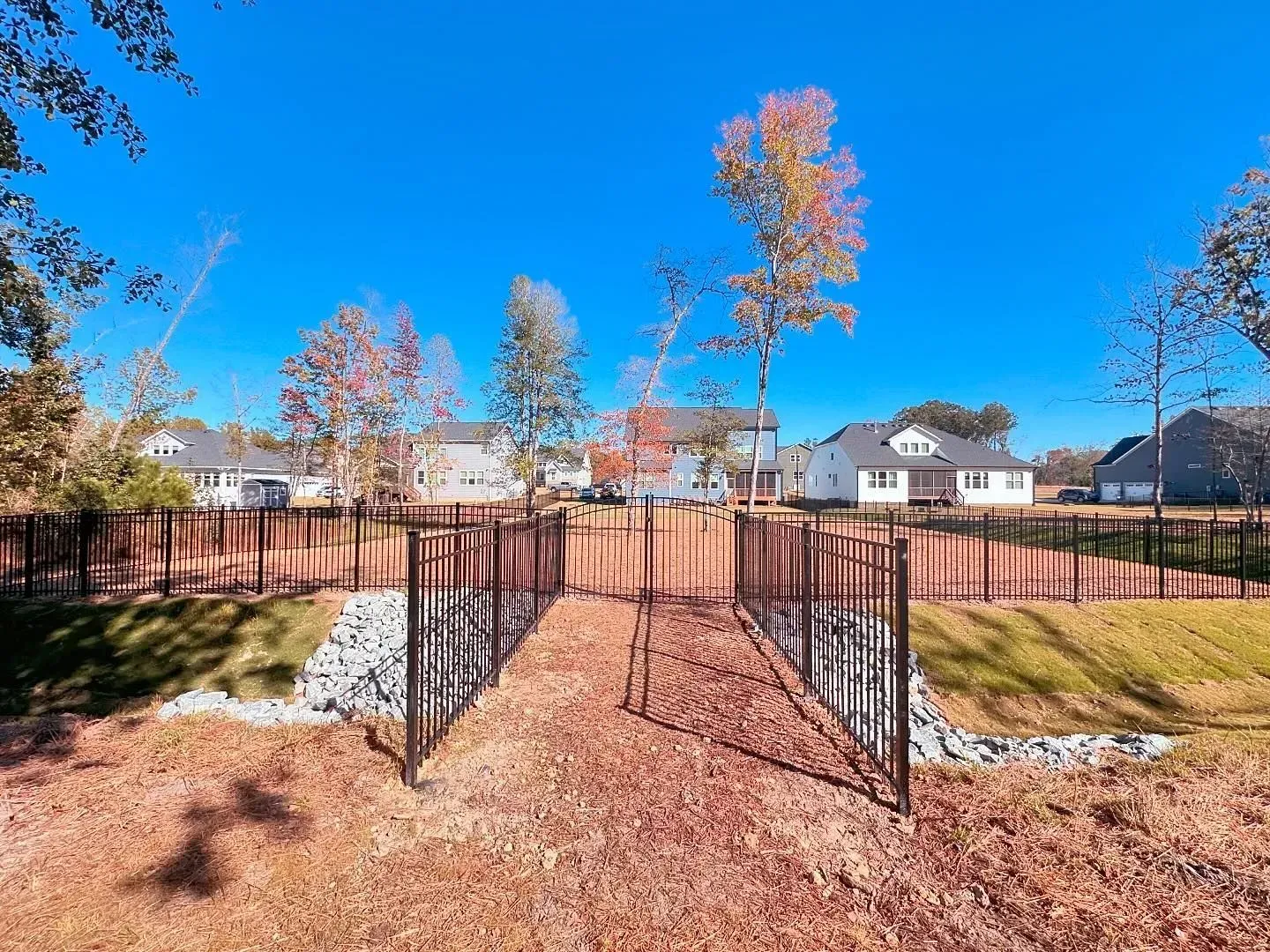 Black metal fence with a gate, houses, and trees under a clear blue sky.
