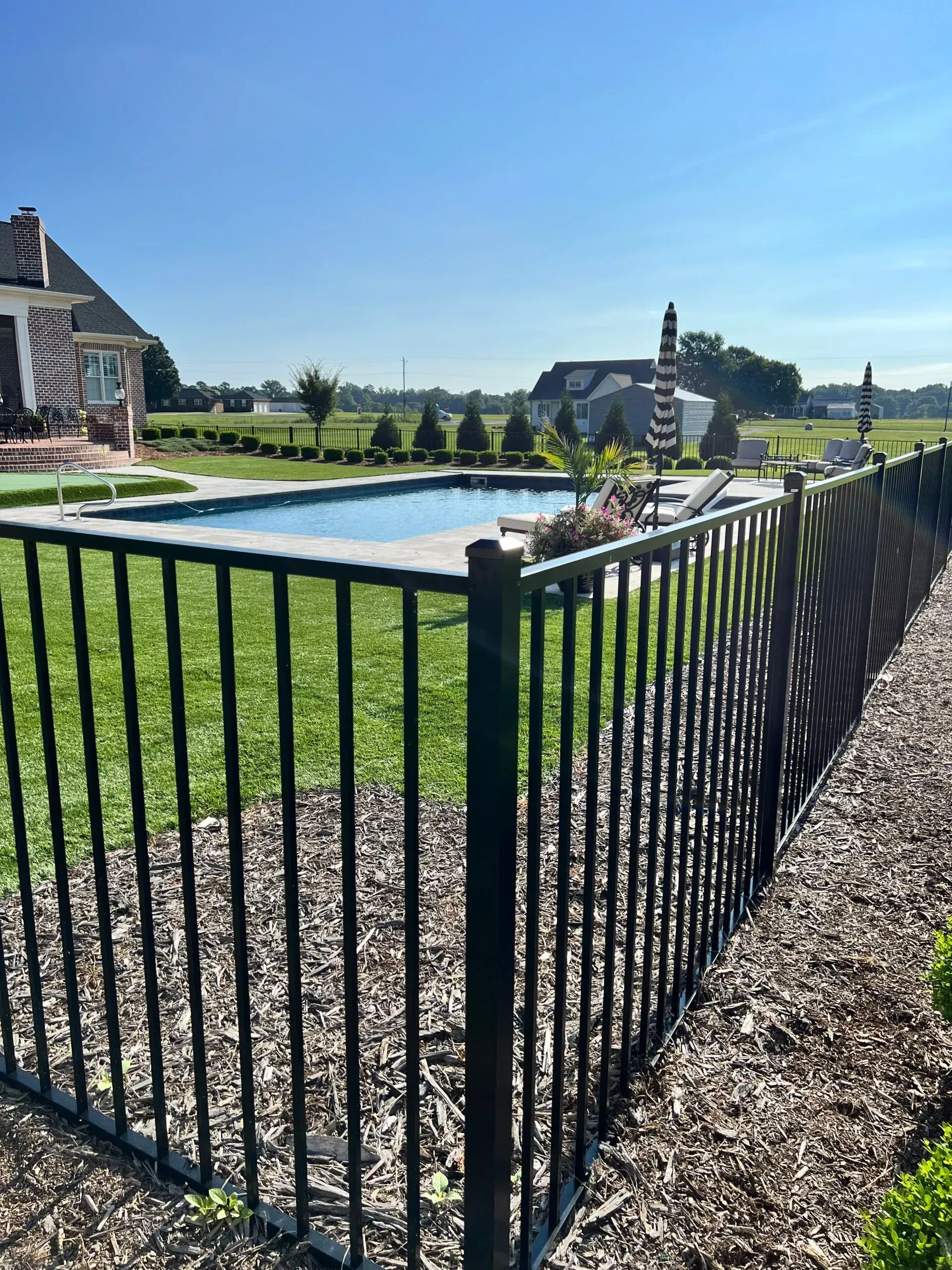 Black fence surrounds a swimming pool on a sunny day; large house and open field in background.