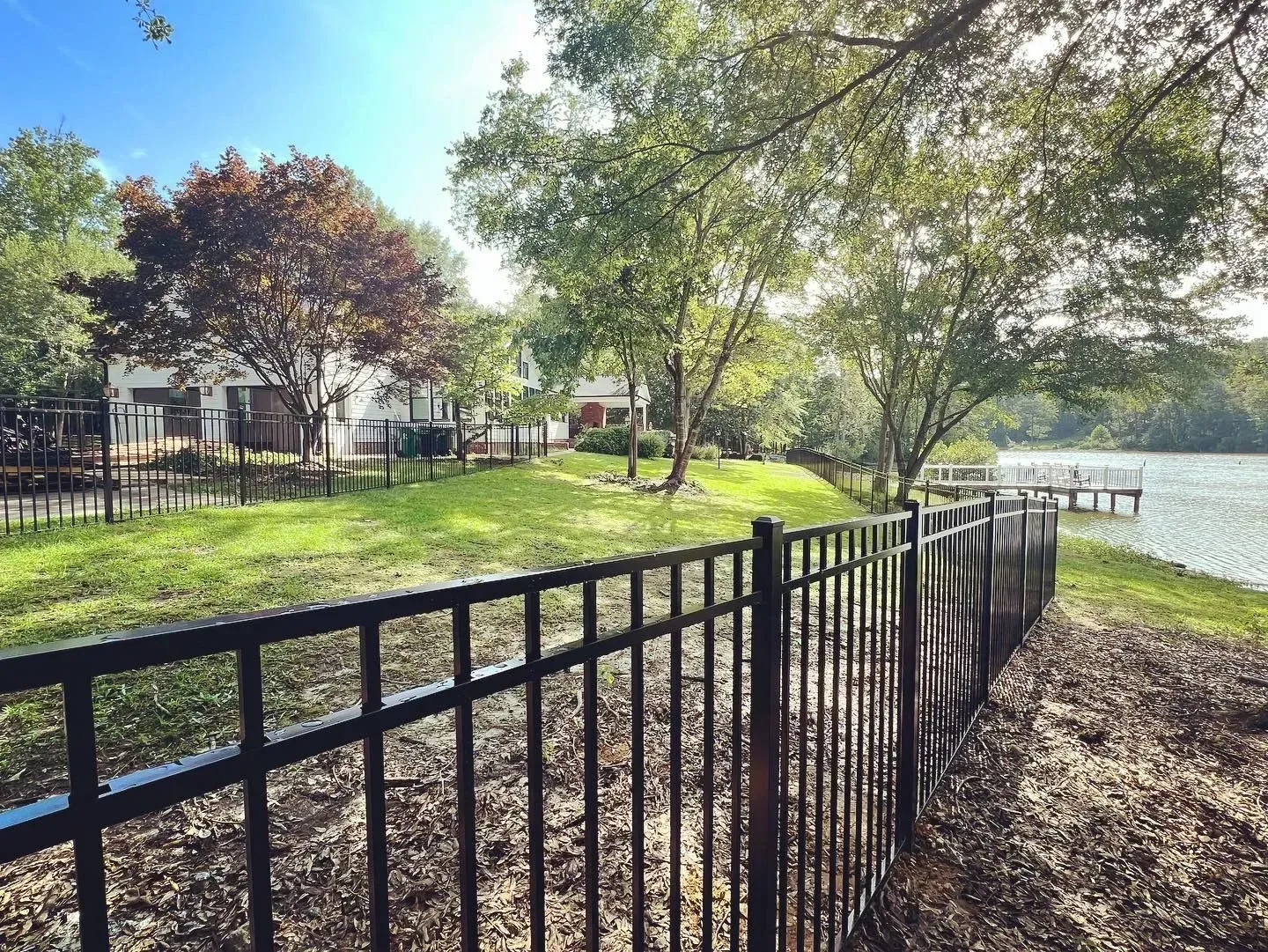 Black fence along grassy bank beside a lake, trees and houses in the background. Sunny day.