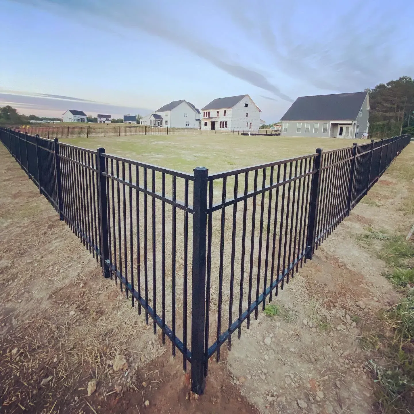 Black metal fence enclosing a large grassy area with several houses in the background under a cloudy sky.